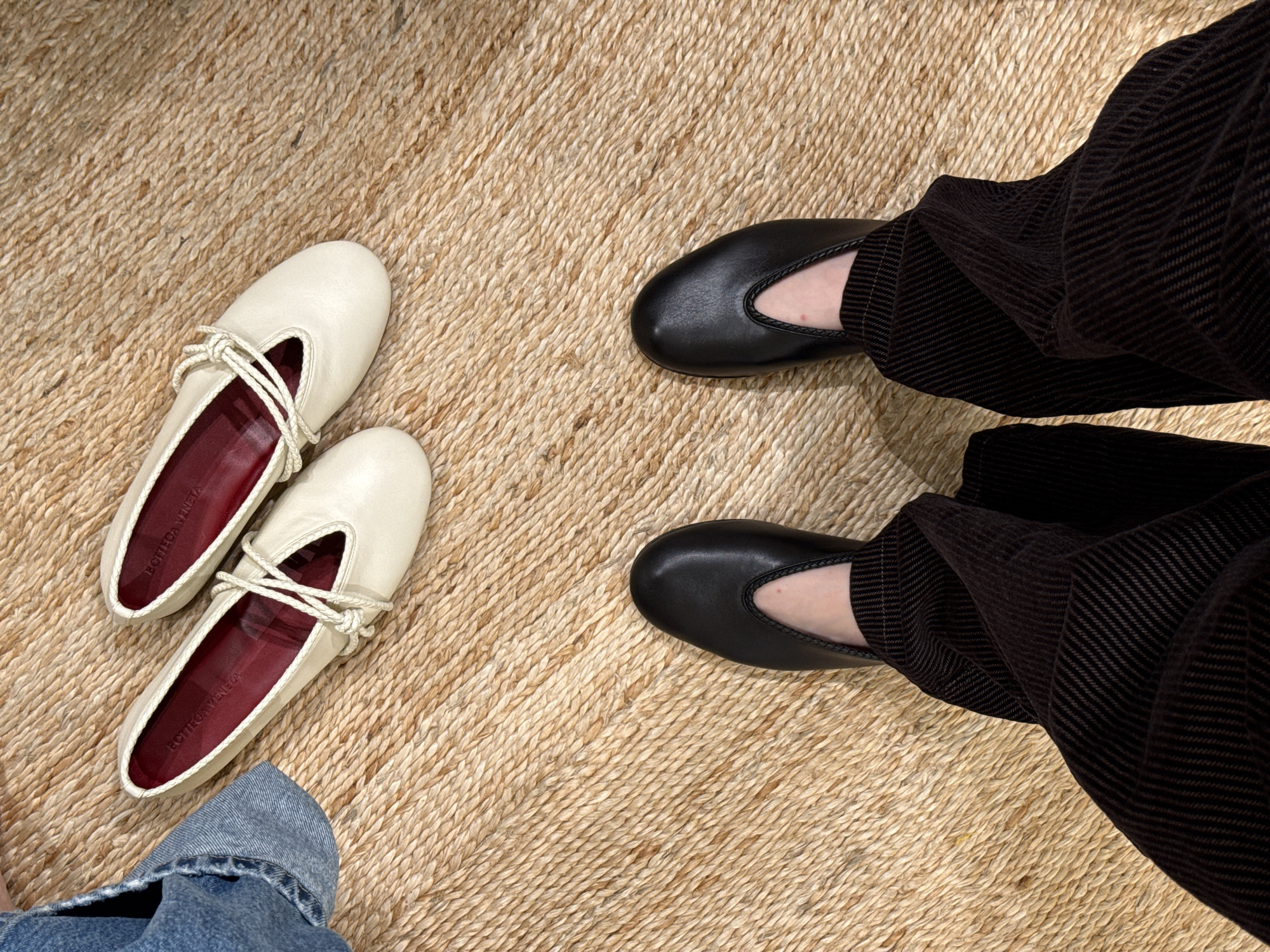 The scene, likely indoors and not specifically identifiable to San Diego, United States of America, features a textural jute rug as the background. A pair of off-white leather ballet flats with maroon lining and intertwined rope detail rest at the left. To the right, a person wears black leather ballet flats with a rounded toe. The person is wearing dark brown, wide-wale corduroy pants. A small section of denim fabric is visible near the lower left corner.