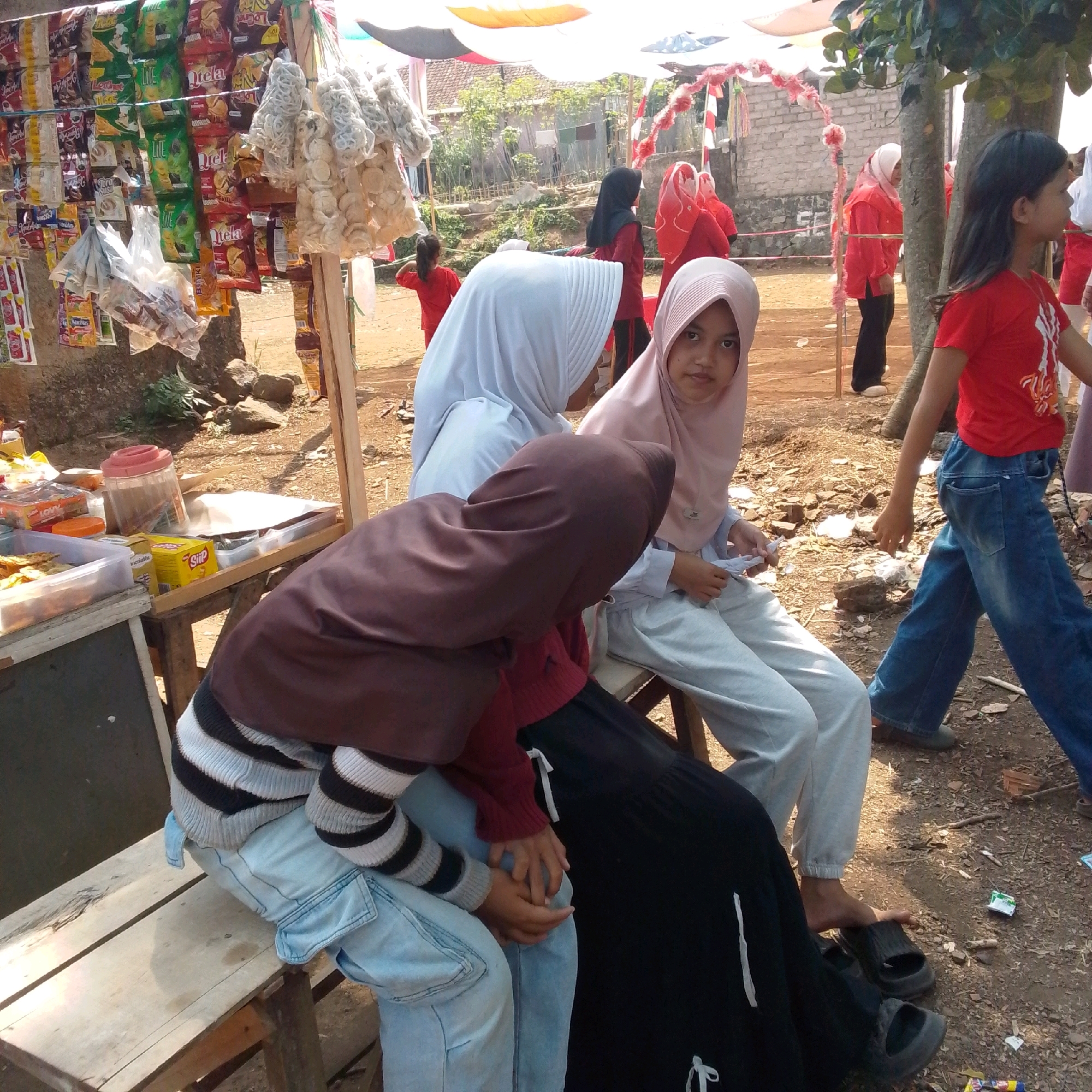 In Cilaku, Indonesia, a group of young people are gathered around a roadside stall. The stall displays various snacks and packaged goods hanging from a wooden frame. Several individuals are wearing hijabs, suggesting a local cultural context. One person is walking away from the stall, wearing a red t-shirt and blue jeans. Another person, also in red, is standing nearby. The ground is dirt and strewn with debris. The overall atmosphere appears to be casual and communal.
