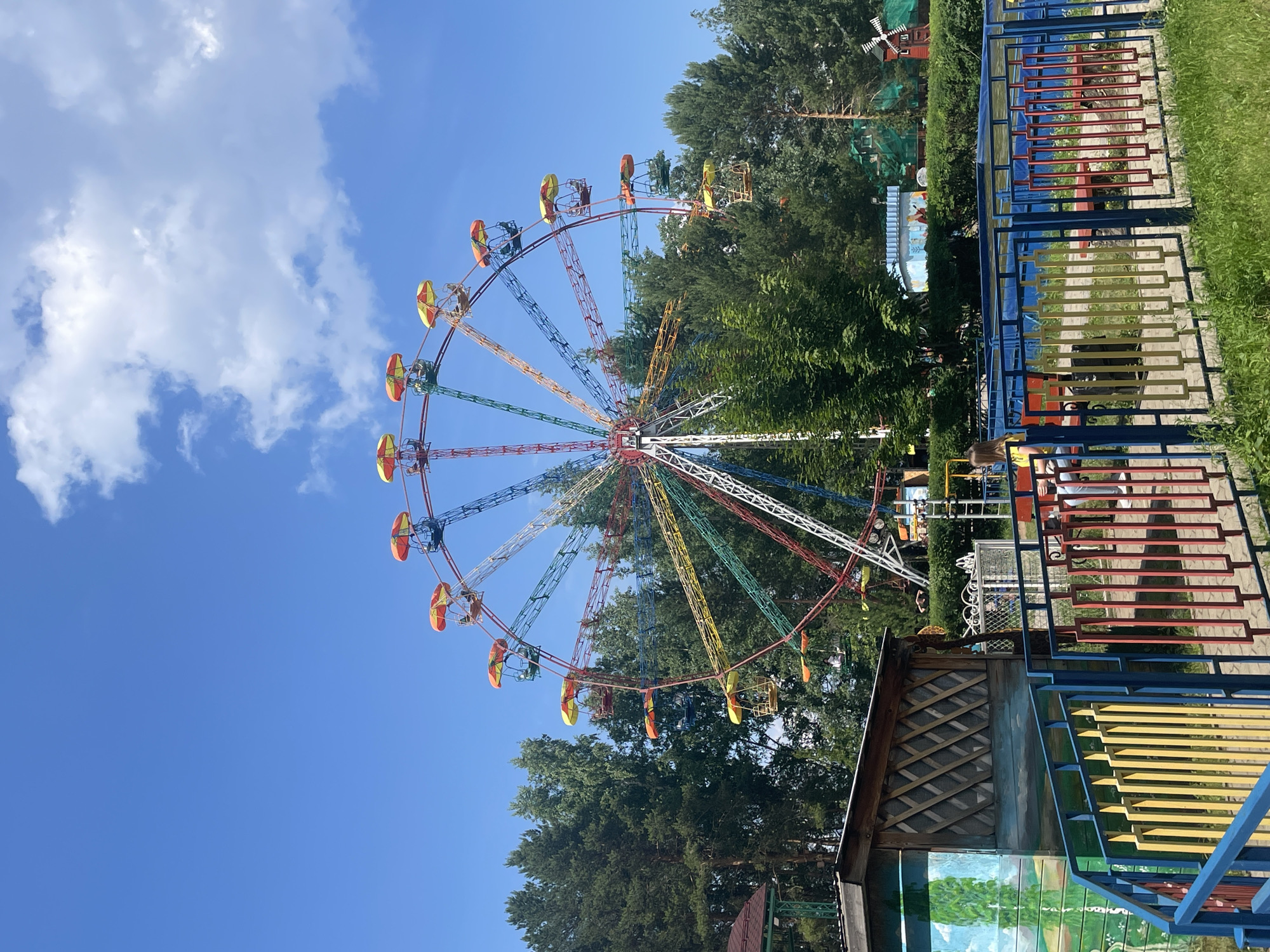 The image shows a Ferris wheel in an amusement park on a clear day. The Ferris wheel is colorful, with red, yellow, green, and blue segments. It has multiple cabins with yellow and orange umbrellas. In the foreground, there is a blue, yellow, and red fence. Behind the fence, there is a small wooden building with a mural on its side. In the background, there are tall green trees. A person wearing a yellow shirt is sitting on a bench in the middle ground. The Ferris wheel is located in the city of Chaikovsky, Russia.