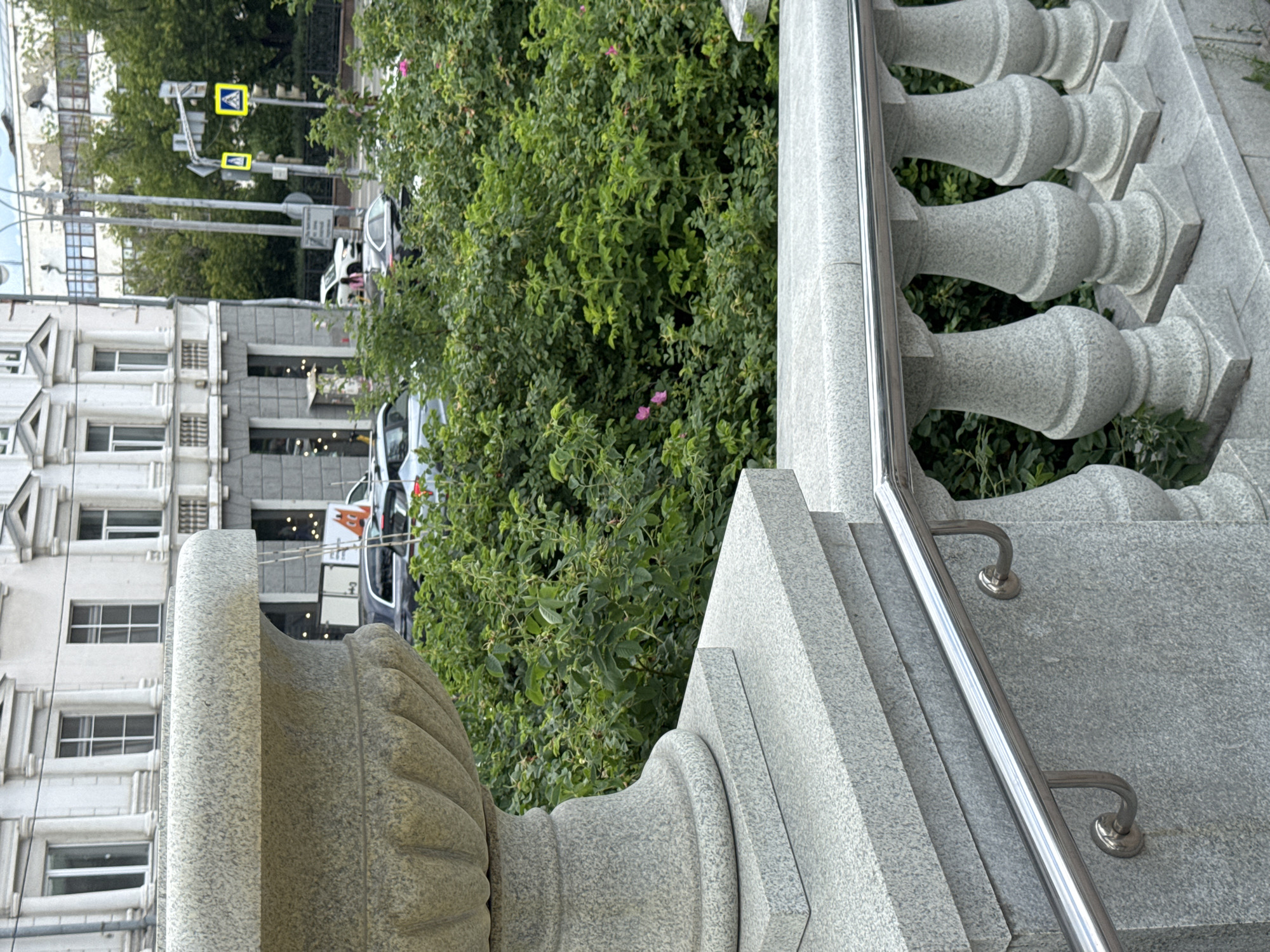 The image captures a scene in Yekaterinburg, Russia, focusing on a stone railing and planter in the foreground. The railing, made of light-colored granite, features ornate balusters and a polished metal handrail. Behind the railing, lush green foliage with scattered pink flowers suggests a garden or park setting. In the background, a multi-story building with classical architectural elements, including columns and arched windows, stands across a street. Traffic lights and yellow warning signs are visible on a traffic pole, indicating a road intersection. Several cars are present on the street, including a white car and a dark-colored sedan. The overall atmosphere suggests an urban environment with elements of classical architecture and green spaces.