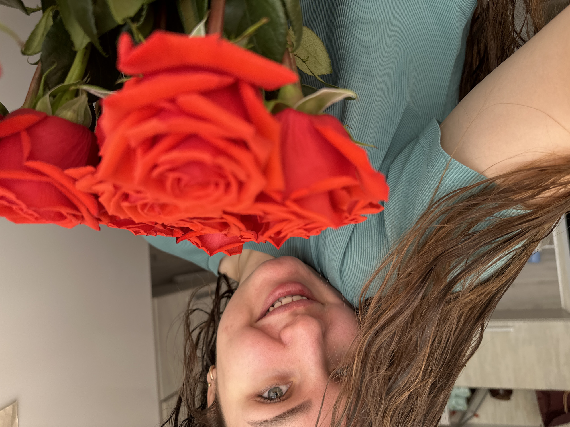 A woman poses for a photo holding red roses. The setting appears to be indoors, possibly in an apartment or home, given the background featuring walls, furniture, and possibly a doorway. The location may be Chaikovsky, Russia, based on the prompt, though there are no readily identifiable markers within the image to confirm this.
