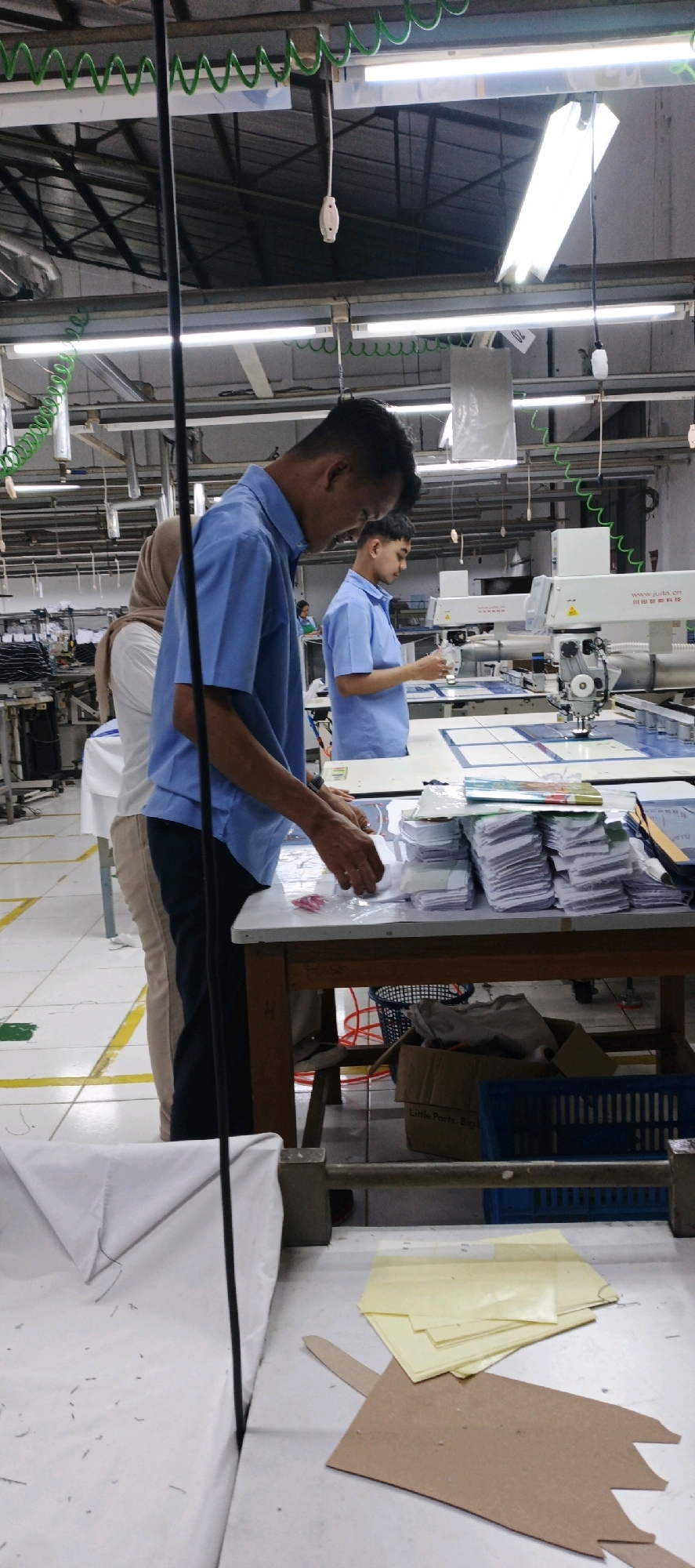 The image depicts an indoor scene within a garment factory in Salatiga, Indonesia. The environment is brightly lit by fluorescent lights hanging from the ceiling. In the foreground, a person wearing a blue short-sleeved shirt and dark pants is working at a table, with their hands engaged in arranging or inspecting small pieces of fabric. Several stacks of folded fabric pieces are visible on the table, along with some sheets of yellow paper and a brown cardboard cutout. A black pole extends vertically through the center of the frame.
In the mid-ground, another person, also in a blue short-sleeved shirt, is standing and working at a sewing machine, which appears to be an automated industrial model. To their left, a person wearing a beige headscarf and a white shirt is standing partially obscured, appearing to be involved in the same production line. The background reveals more industrial equipment, including sewing machines and tables, suggesting an active production floor. The floor is tiled in a light color with yellow safety markings. A blue plastic basket is visible in the lower right corner, and a cardboard box with some items inside is near it. The overall impression is a busy, functional manufacturing environment.