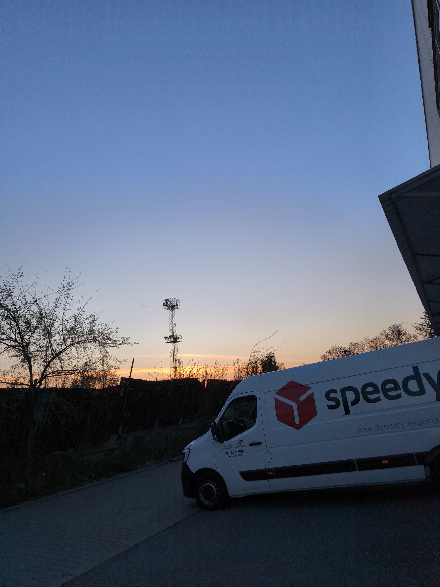 The image captures an outdoor scene at dusk, characterized by the warm hues of a sunset blending into a deep blue sky. A white delivery van, prominent in the lower right and center, is the main subject. The van, likely a Renault Master, features the "Speedy" logo in large black letters with a red cube graphic on its side. Below the brand name, the slogan "Your delivery experts" is visible. Additional text on the driver's side door includes "MAPPEAOT GLOBPOST", a phone number "0 7001 7001", and the website "www.speedy.bg".
The van is parked on a dark paved surface, with an adjacent asphalt road. To the left, silhouetted bare trees and a dark fence line the horizon, where the sun casts an orange and yellow glow. A tall metal lattice tower, equipped with what appear to be lights or communication dishes, stands in the mid-ground against the sunset. A faint, thin white contrail stretches horizontally across the sky, slightly above the tower. On the right, the lower portion of a building or large structure with an angled grey roof overhang is visible, suggesting a commercial or industrial setting, possibly a warehouse or depot. The overall atmosphere is tranquil, marking the transition from day to night in what is identified as Burgas, Bulgaria. No people or active events are depicted.