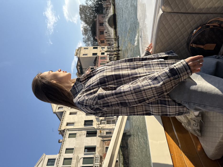 A young adult female is depicted standing in profile, facing right, on a boat in a Venetian canal. She wears a brown, beige, and dark blue plaid long-sleeved shirt over a white undershirt and light blue denim jeans. A silver-toned ring is visible on her left ring finger. Her brown hair is down, parted on the side, and swept back. The boat features light-colored, quilted seating and wooden trim. A brown, monogrammed Louis Vuitton 'Speedy' or similar style bag with a rounded shape and leather handles rests on the seat to her right.
The background reveals typical Venetian architecture: multi-story buildings of varying heights and colors (cream, beige, pink, brown) line the canal. Windows and a balcony are visible on the buildings to the left. In the mid-ground, a stone arch bridge spans the canal, with additional buildings and trees beyond it. The canal water exhibits ripples. The sky above is clear blue with some wispy white clouds. The scene is illuminated by strong sunlight, casting distinct shadows. The overall environment identifies the location as a canal in Venice, Italy.