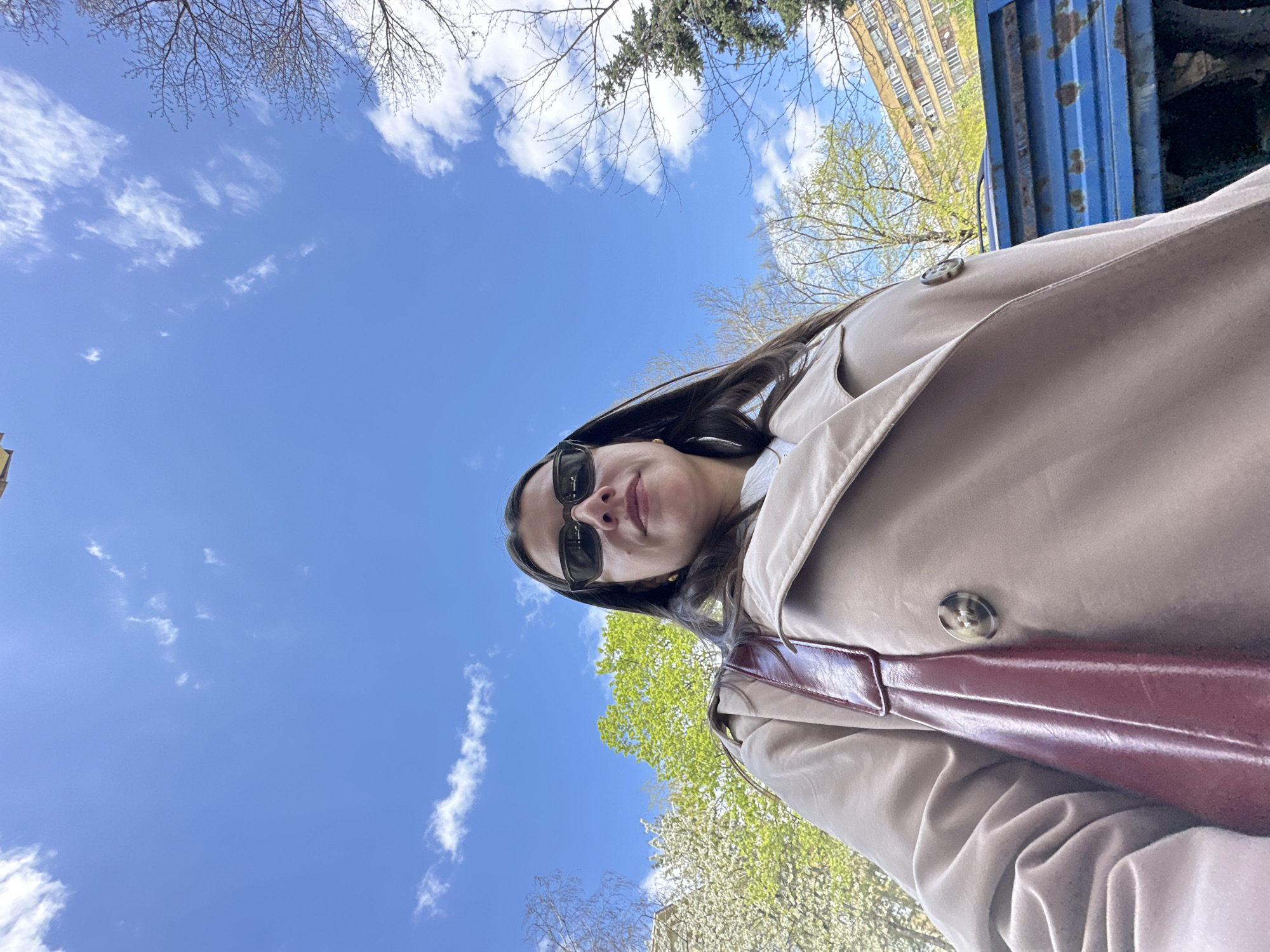 The image captures a low-angle, upward-facing shot of a young woman outdoors in Sofia, Bulgaria, on a bright, clear day, likely in spring. The woman is positioned in the lower-right portion of the frame, looking down towards the camera with a subtle smile. She has long dark hair and is wearing dark sunglasses, a light-colored (beige or tan) trench coat with a prominent button, and a dark red or burgundy shoulder bag slung across her body.
The sky, a vibrant blue with scattered white clouds, fills most of the upper frame. Surrounding the sky and the woman are various trees, some with bare, intricate branches suggesting early spring or deciduous varieties, while others are adorned with fresh, bright green foliage. Evergreen trees are also visible in the background. In the upper-left background, a multi-story, light-colored building with many windows, characteristic of an apartment block, is partially visible. To the upper right, a dark blue, heavily rusted corrugated metal structure, possibly an urban fixture or container, adds an industrial touch to the scene. The bright, natural lighting indicates it is daytime, with pleasant weather conditions. No visible text is present.