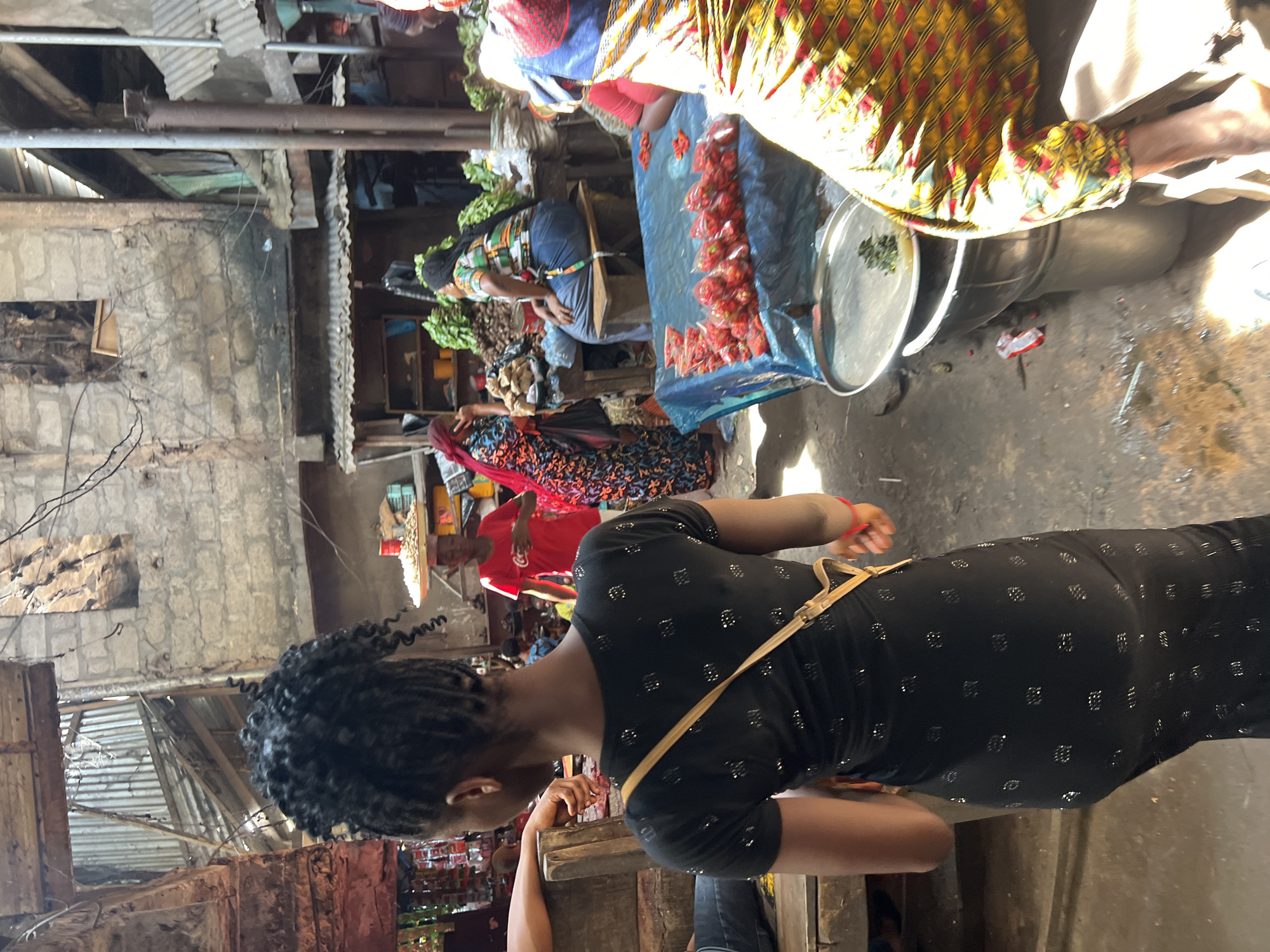 This image depicts a bustling market scene in Nkpor, Nigeria. The foreground is dominated by a young woman wearing a black, sparkly dress and a tan crossbody strap. Her hair is styled in intricate braids, and she is facing away from the camera, looking into the market. To her left, a man's arm rests on a wooden structure.
The market environment is characterized by an open-air structure with corrugated metal roofing and exposed wooden beams. The background reveals stalls with various goods, including piles of vegetables and numerous small bags of red peppers laid out on a blue tarp. Several other individuals are visible in the midground and background, engaged in shopping or selling. One person wears a red t-shirt, another a brightly colored patterned dress, and others are partially obscured. The ground appears to be dirt or concrete, with some wet patches and debris. The overall impression is one of a vibrant and active commercial hub.