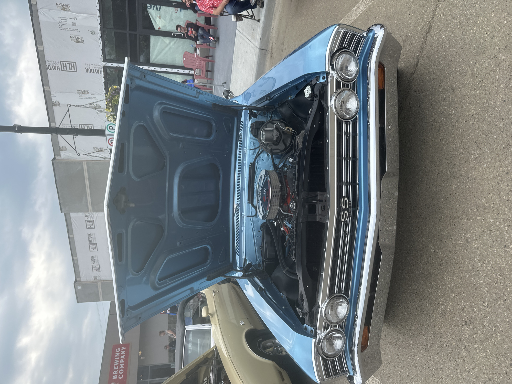 This media file captures a close-up, high-angle shot of the front of a light blue classic car, likely a Chevrolet Chevelle SS, with its hood open. The car is parked on a paved surface, possibly a street or parking lot, with a building and some people visible in the background.
The car's engine is exposed, revealing a chrome air cleaner and various engine components. The front grille, headlights, and chrome bumper are highly polished. The "SS" emblem is clearly visible on the grille. To the left of the classic car, the front of a lighter-colored vintage car is also partially visible.
In the background, a building with large windows is under construction, covered in white sheeting that displays "HLH HAYDUK LUMBER AND HARDWARE" and "TITAN" text. A red sign with white lettering reads "BREWING COMPANY". Several people are seen standing and sitting in the background, some on chairs, suggesting a car show or community event. The lighting suggests it is daytime, with overcast or partly cloudy skies. The overall impression is of a car enthusiast gathering or a local event showcasing vintage automobiles.
