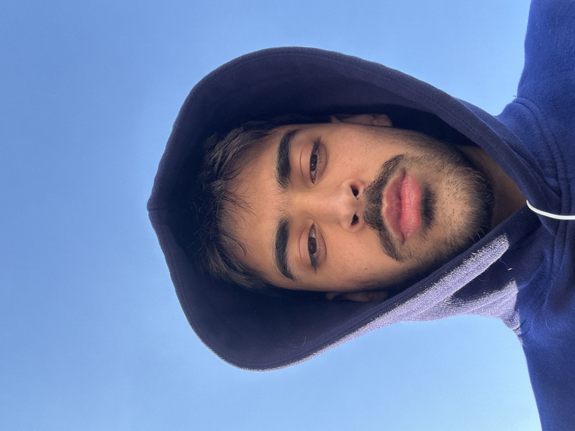 A man with a dark beard and mustache is wearing a blue hooded sweatshirt. The hood is pulled up over his head. The sky is blue and clear in the background. The image appears to have been taken from a low angle, looking up at the man. There is no information in the provided image to suggest that the image was taken in the city of Mirandela, Portugal.
