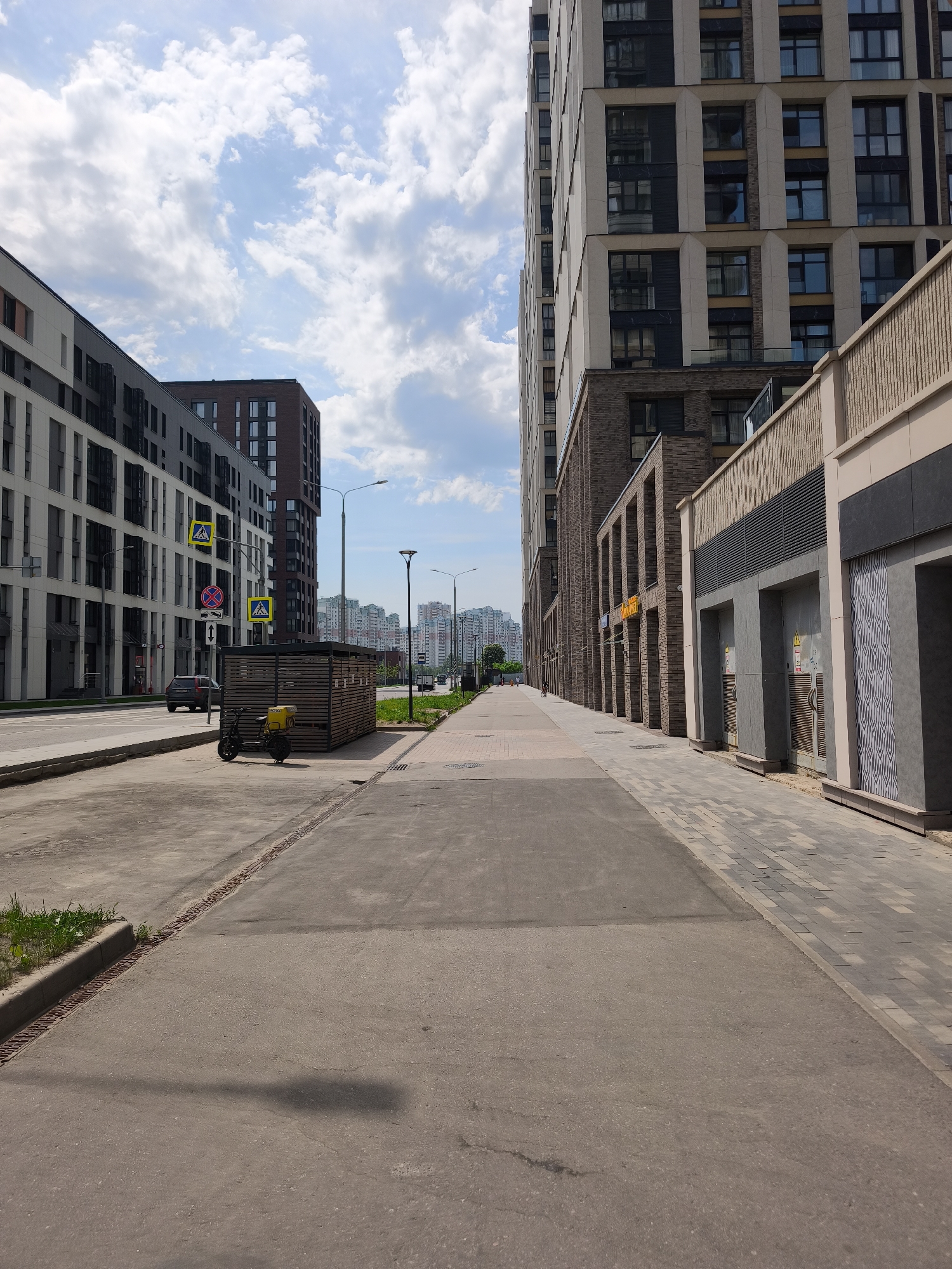 The image depicts a wide pedestrian walkway in a modern urban setting, possibly Moscow, Russia, based on the architectural style and signage. The walkway is paved with concrete and leads toward distant high-rise buildings, suggesting a residential or mixed-use area.
On the left, a street runs parallel with pedestrian crossings and a black delivery motorbike parked in front of a wooden structure. Modern apartment buildings with dark and light facades are visible along the street.
On the right side of the walkway, the facade of a multi-story building is visible, with a combination of brick, beige stone, and architectural details. There are commercial spaces or entrances along the base of the building.
The sky is partly cloudy, casting shadows on the walkway, suggesting it is daytime. There are streetlights along the pedestrian path and the roadway. The overall impression is a modern, well-planned urban area.