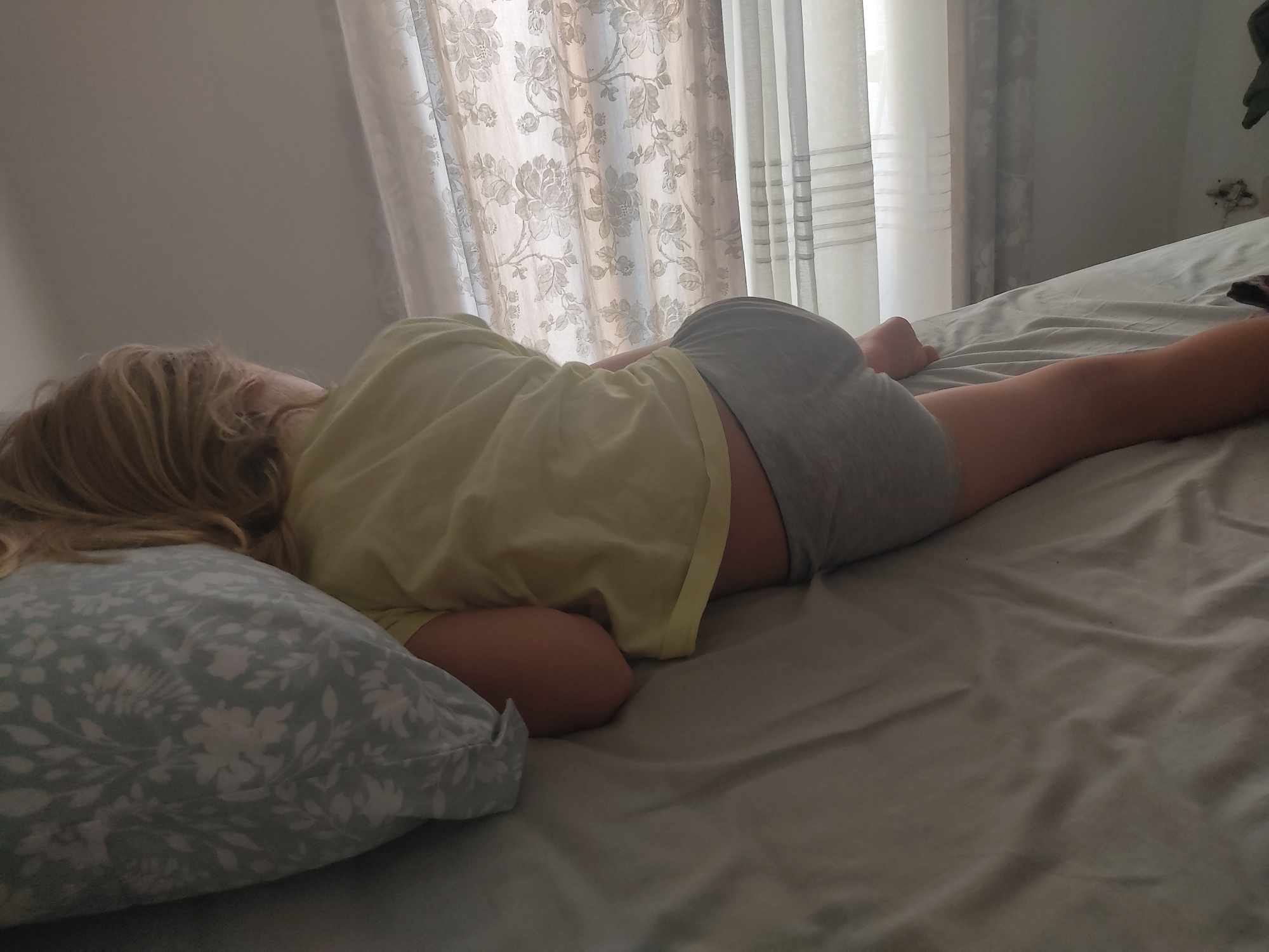 A person with blonde hair is sleeping on their side on a bed. They are wearing a yellow t-shirt and grey shorts. The bed has a light grey duvet and a light blue pillow with a white floral pattern. In the background, there are sheer curtains with a floral pattern. The lighting suggests it is daytime. The image was taken in Blanes, Spain.