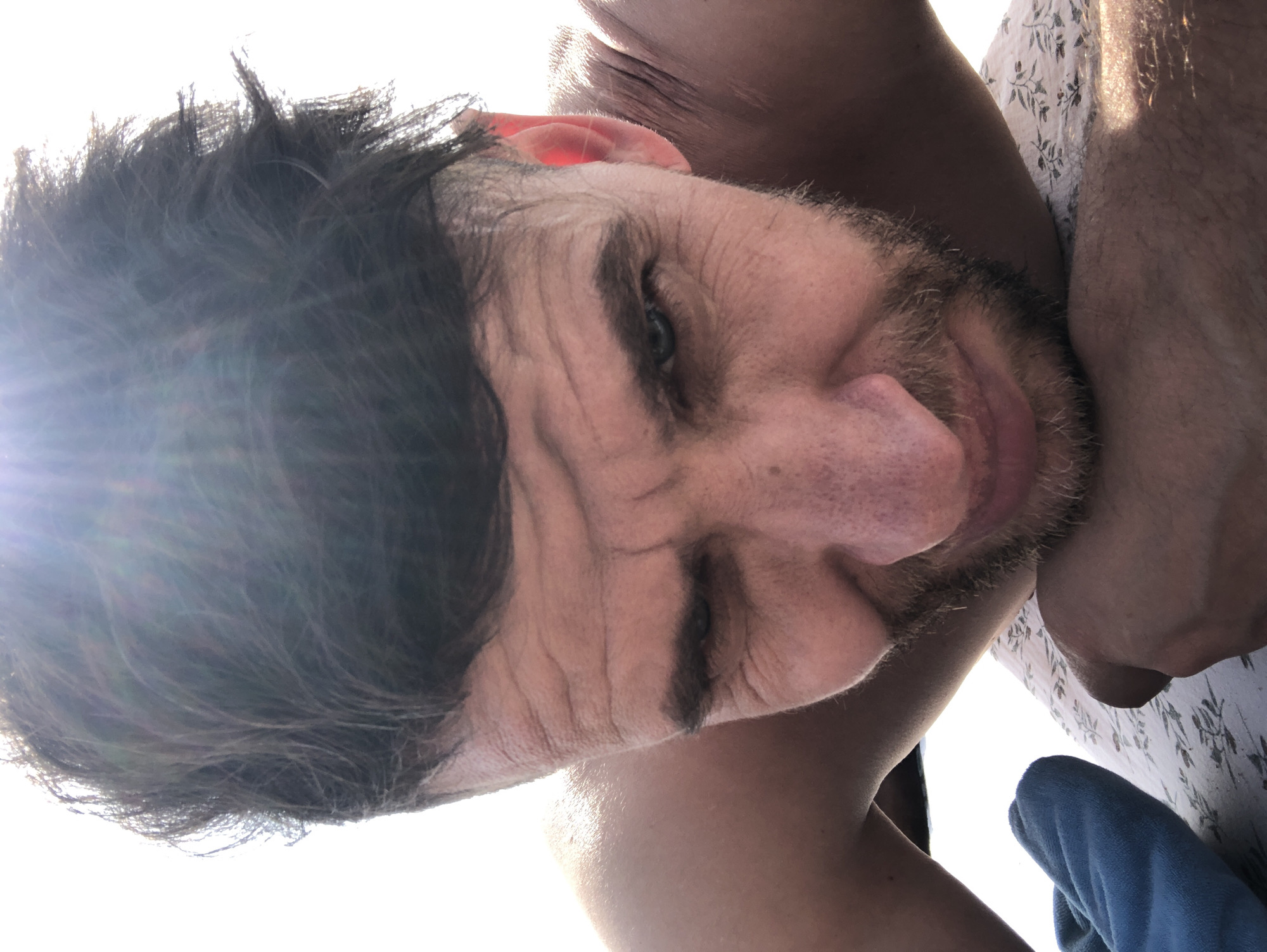 A close-up, low-angle shot features a man with dark, disheveled hair and a short beard, appearing to be in his late 30s to 40s. He is looking downwards, with his blue-green eyes partially visible and prominent wrinkles on his forehead and around his eyes. His skin is a fair to medium complexion, with visible pores on his nose and cheeks. The upper part of his bare shoulders and arms are visible, suggesting he is shirtless. Strong sunlight from the upper left casts a bright glare and creates a lens flare effect. The background is overexposed and appears pure white, obscuring any discernible environmental details. Beneath him, a white fabric with a subtle, repeating dark leaf or floral pattern is visible, and to the left, a folded piece of bright blue fabric is present. The specific location in Sestroretsk, Russia, cannot be determined from the visual information provided in the image.