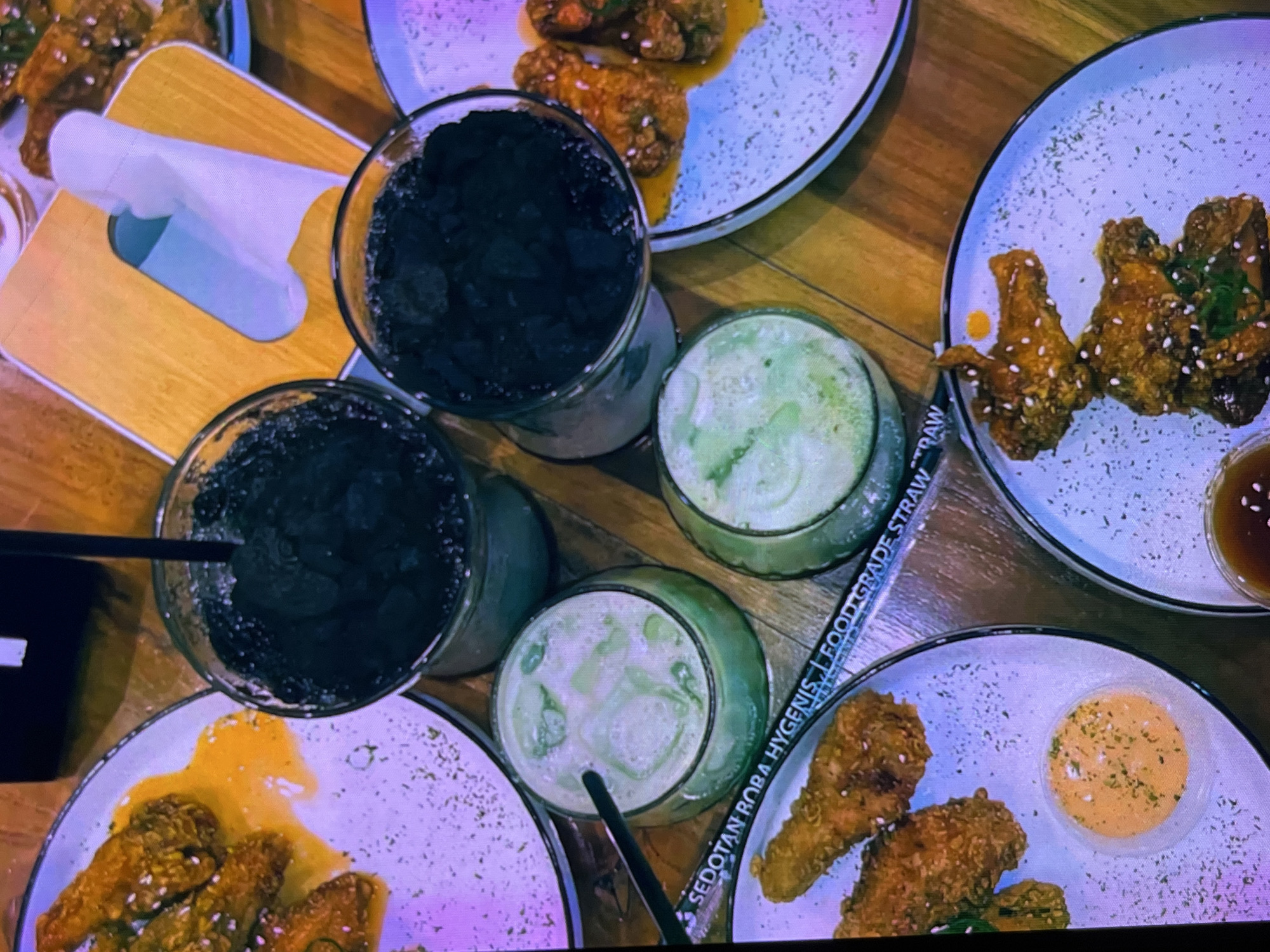 The image shows a table laden with food and drinks, suggesting a meal at a restaurant or cafe. Plates of fried chicken wings, some with a yellow sauce, are visible. There are also several glasses containing beverages; two appear to be dark-colored and possibly iced, while two others are a pale green color and also appear to be iced. A black straw is placed in one of the green beverages. A white paper wrapper with the words "SEDOTAN BOBA HYGENIS FOOD GRADE STRAW" is visible in the foreground. The table surface appears to be made of wood. The lighting is dim, with a purple tint. There are no people clearly visible in the image. The setting is consistent with a dining establishment. The location in Cadek, Indonesia, is not explicitly depicted or deducible from the visual information provided in the image.