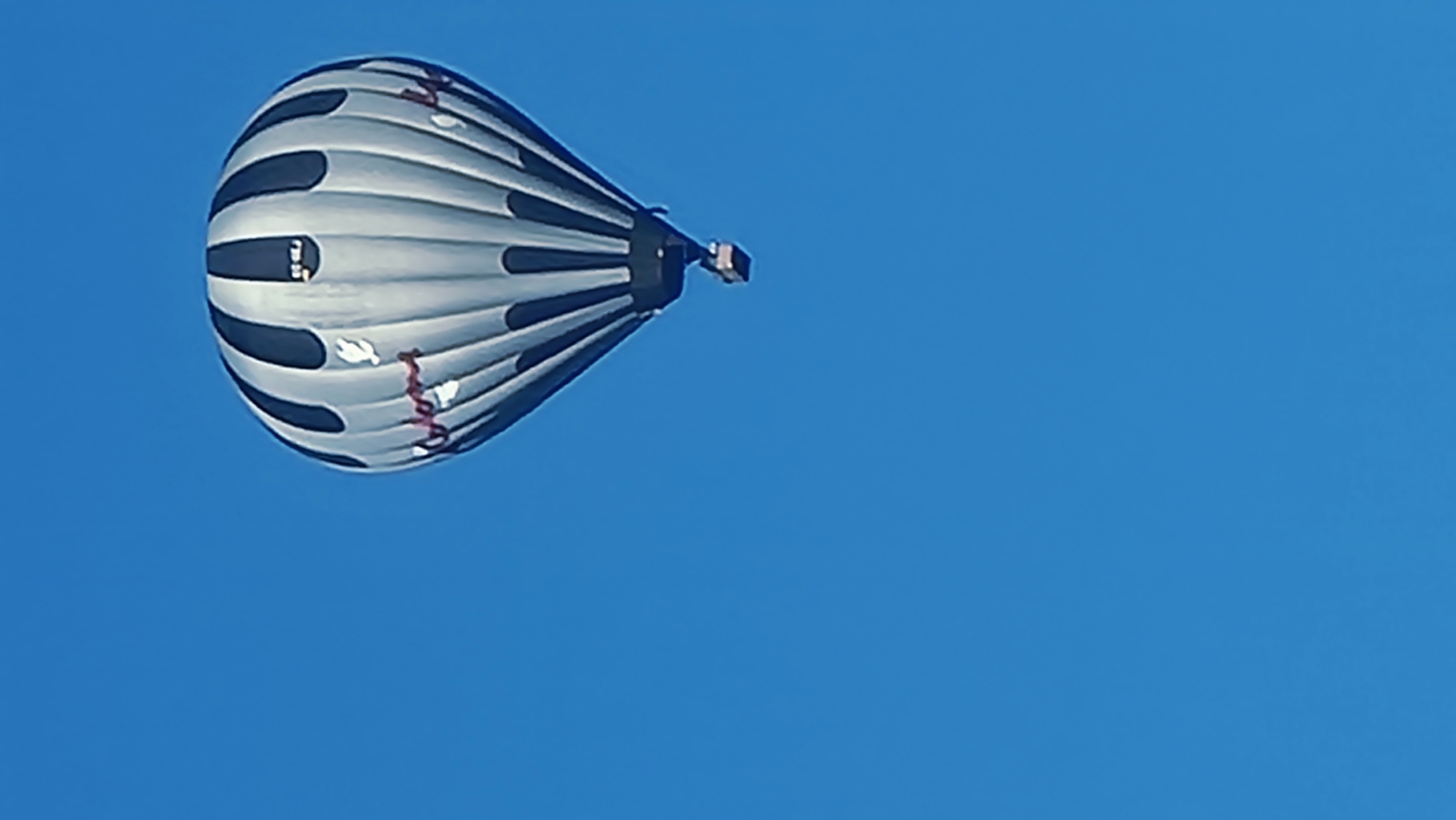 A hot air balloon with black and white vertical stripes is suspended against a clear blue sky. The balloon features some red lettering and white graphic elements. A dark basket hangs beneath it. The image was captured from a low angle, looking up at the balloon. The location is Sant Joan Les Fonts, Spain.