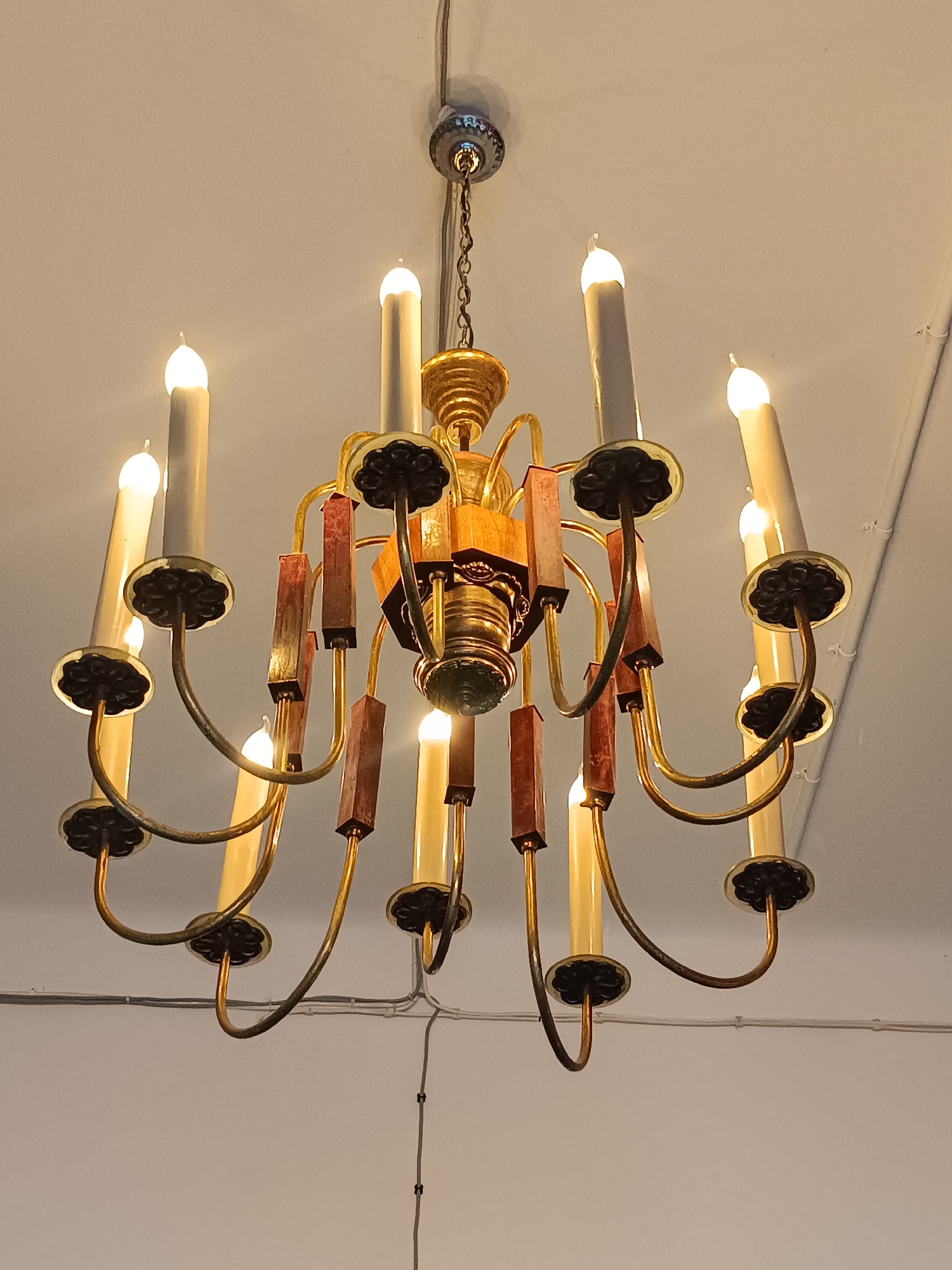 A brass and wood chandelier with twelve candle-shaped lights is hanging from a white ceiling. The chandelier has a central wooden element and brass arms that curve outwards to hold the lights. The lights are a warm, off-white color and are all illuminated. Some of the arms have rectangular wooden accents. The image appears to be taken indoors. No specific location in Riga, Latvia, can be determined from the image.