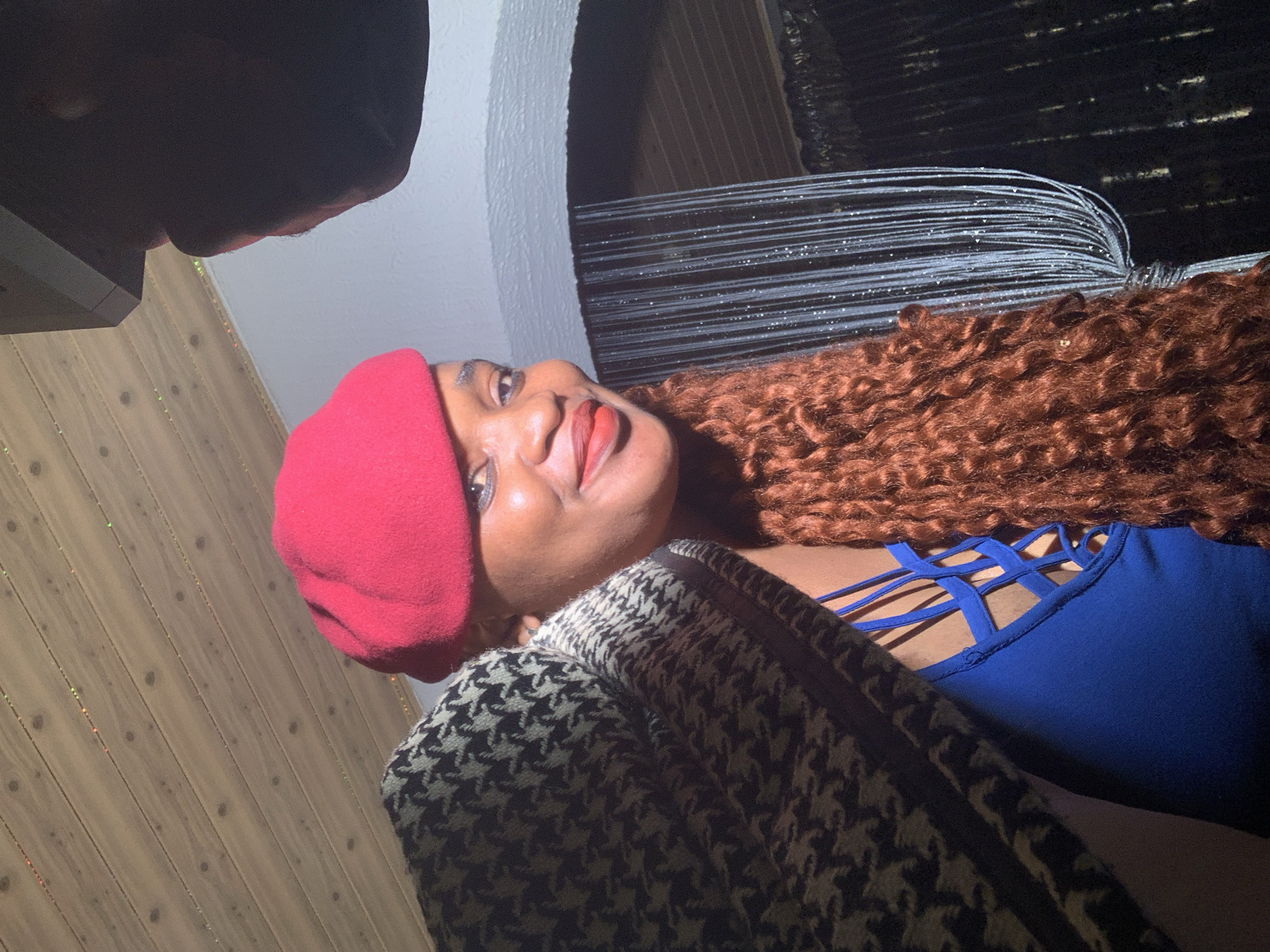 An indoor portrait features a woman smiling broadly with her head tilted upwards and to the side, looking off-camera. She has dark skin, full red lips, and long, voluminous, curly reddish-brown hair flowing over her right shoulder. She wears a striking red beret and a vibrant blue top with a distinctive criss-cross strap design at the neckline. A black and white houndstooth-patterned jacket or shawl is draped over her shoulders.
In the upper left corner, a dark, indistinct shape is partially visible, possibly another person's head or an object close to the camera. The background on the left consists of vertical wooden paneling adorned with small, colorful, twinkling lights. To the right, a white archway frames a darker area, beyond which multiple thin, reflective, silvery strands or filaments are visible, creating a textured backdrop.
The scene is lit by artificial light, suggesting an evening or nighttime setting. The woman's joyful expression indicates a happy or relaxed mood. There is no discernible text in the image. The setting is Barakin Akawo, Nigeria.