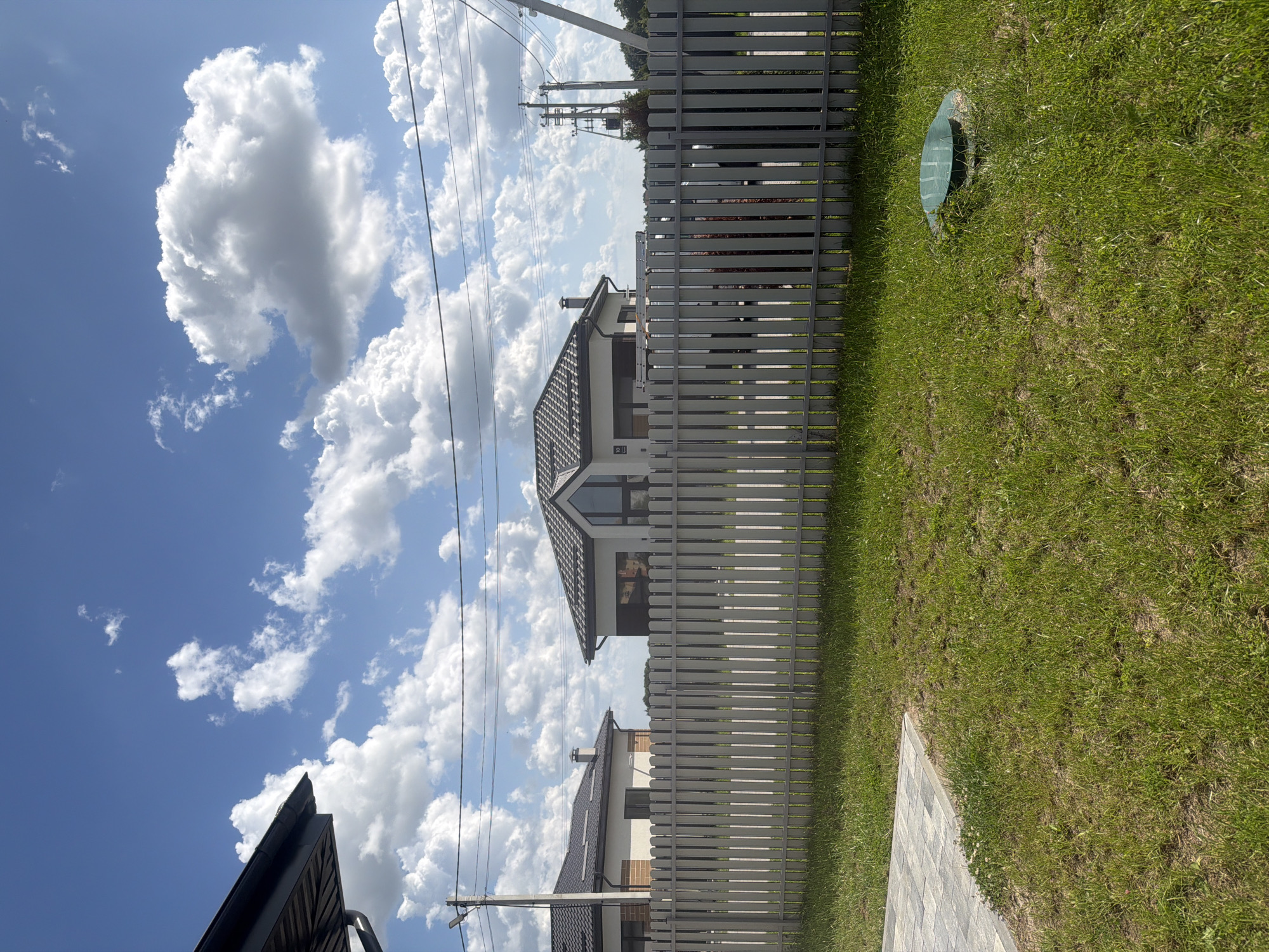 The image shows a residential scene in Golubevo, Russia, on a sunny day with scattered clouds. In the foreground, a light-colored wooden fence runs horizontally across the frame, dividing a manicured lawn from the view beyond. The lawn is green and well-maintained, with a section of grey interlocking pavers visible in the lower left corner. A green septic tank cover is set into the grass on the right side of the lawn.
Behind the fence, two modern houses with dark tiled roofs are visible. The house on the left has a light-colored facade with brick accents and a prominent chimney. The house on the right features a larger, more angular design with a large, arched window. Electrical poles and overhead wires are present in the background, indicating the presence of infrastructure. The sky is a clear blue with voluminous white clouds. No people are present in the image.