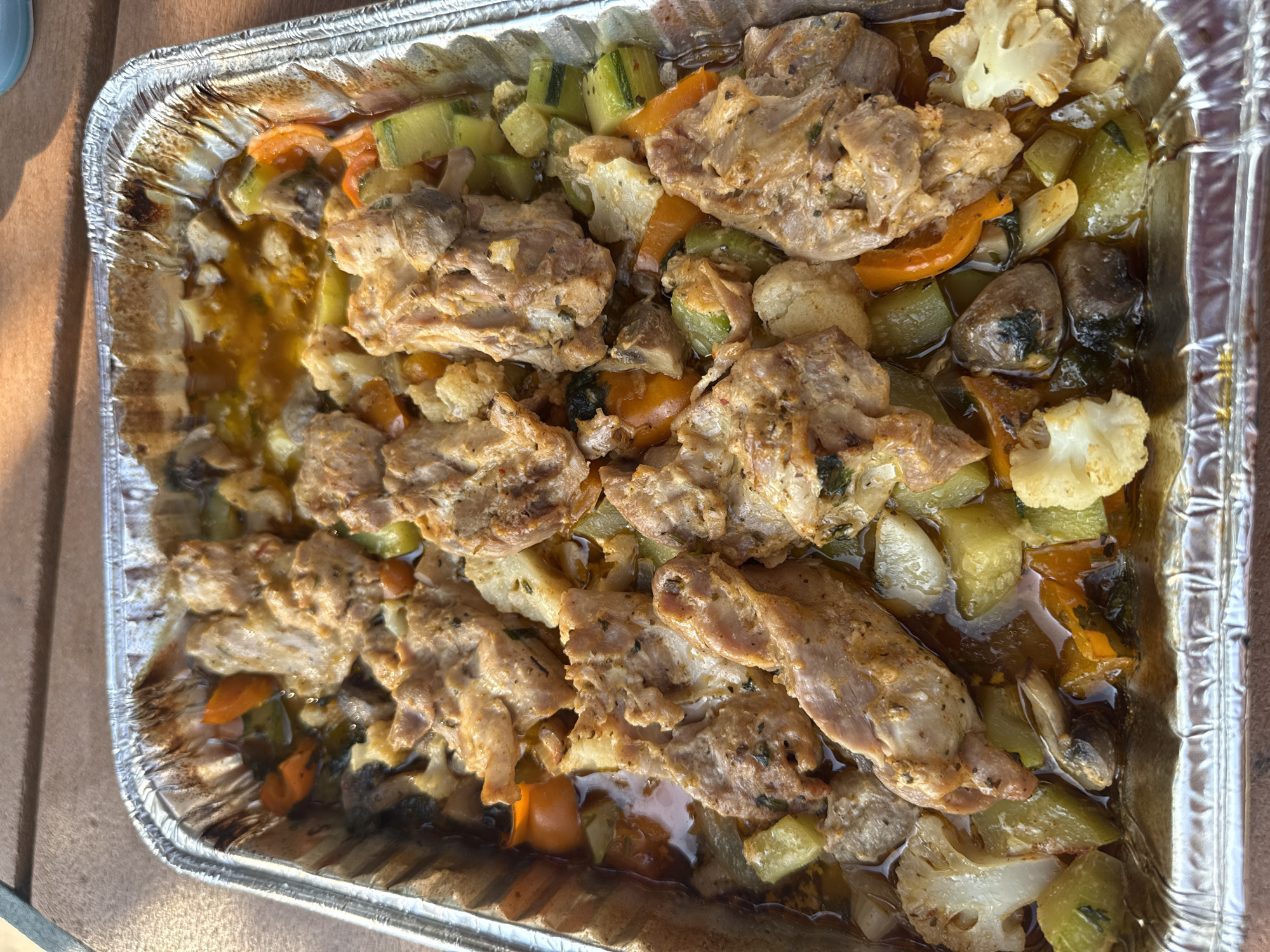 A foil pan filled with roasted meat and vegetables sits on a wooden table. The meat appears to be chicken or pork, cut into bite-sized pieces and seasoned. Among the vegetables are cauliflower florets, sliced zucchini, carrots, and mushrooms. The dish is coated in a light sauce or juices, indicating it was likely baked or roasted. The lighting suggests it is daytime, with sunlight casting shadows. The background is blurred but appears to be an outdoor setting, possibly a backyard or patio. The image was taken in Allen Township, United States of America.