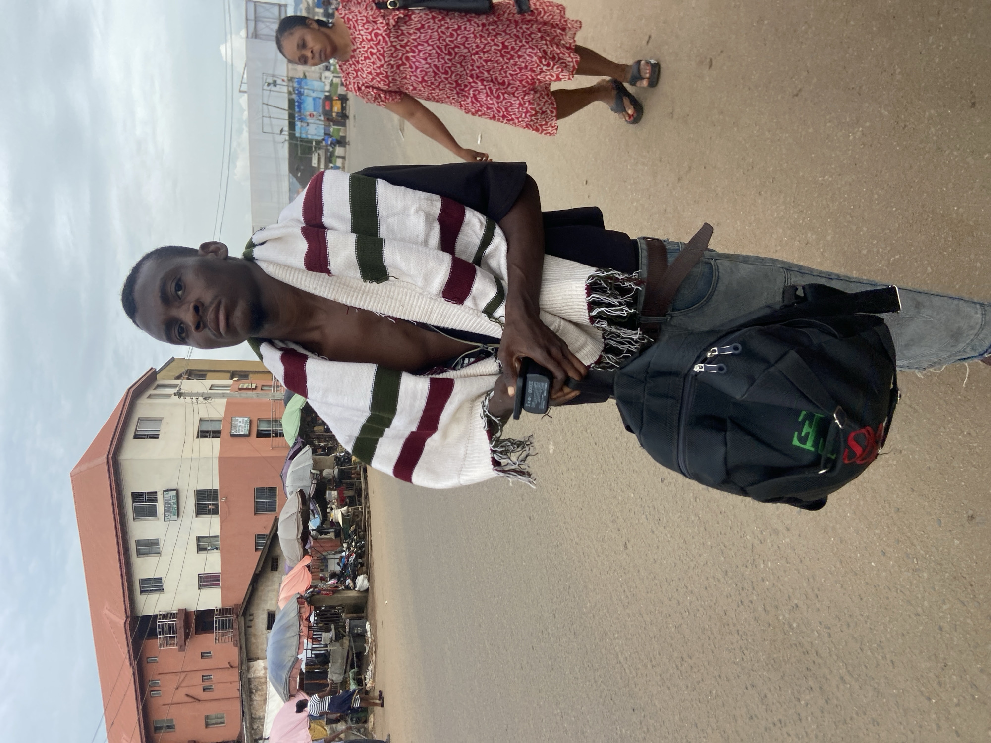 In Nkpor, Nigeria, a young Black man stands in the foreground, facing the camera, with a multi-colored striped scarf draped around his neck and shoulders. He is wearing a black t-shirt, blue jeans, and a dark belt. A black backpack hangs from his left shoulder, and in his hands, he holds a black electronic device. Behind him, a market scene unfolds with various stalls and umbrellas. To the right, a Black woman in a red and white patterned dress walks away from the camera, wearing dark sandals. The background features a multi-story building with a red roof and cream and orange facades, bearing signage for "Summerhill Hospital." The sky is overcast, and the scene appears to be on a paved road.