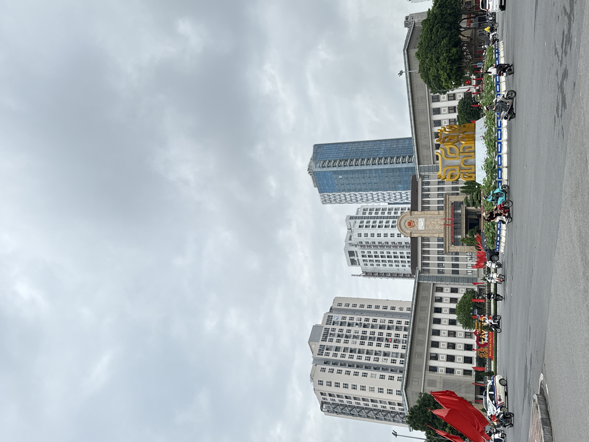 The image depicts a street scene in Hà Nội, Vietnam. A row of parked motorcycles and vehicles line the right edge of the frame. In the background, several tall buildings are visible, including a glass skyscraper and two high-rise apartment buildings with white facades. A governmental building with a golden decorative sign and a clock on its façade is present. Red flags are positioned near the street level. The sky is overcast, covered with gray clouds.