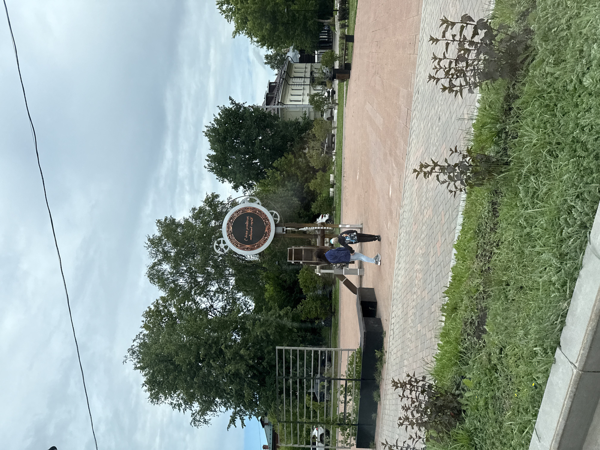 The image depicts a public square in the city of Perm, Russia, on an overcast day. In the foreground, there is a paved walkway and a grassy area with low-lying plants. Two people, an adult and a child, are standing near a decorative circular structure with gears and text in Russian. The text is partially obscured but appears to be a slogan or motto. Behind them, there are trees, buildings, and a metal structure that looks like a trellis or decorative fence. Cars are visible parked in the background, and streetlights are present. The overall impression is that of a well-maintained urban public space.