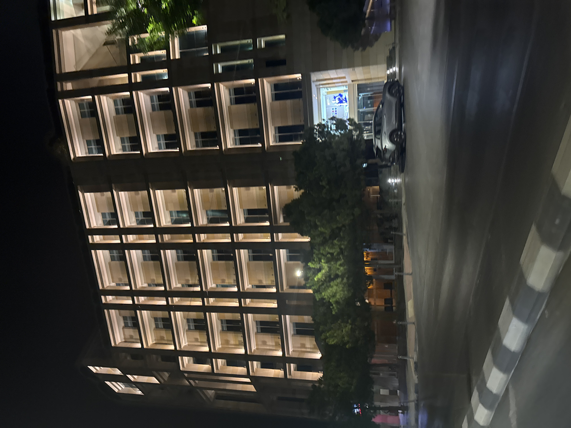 The image shows a modern, multi-story building illuminated at night, with trees lining the street in front of it. The building features a repetitive architectural design with balconies and numerous windows. A silver SUV is parked in front of the building's entrance, which is lit with bright lights and displays a large screen or advertisement. The street in the foreground is wet, suggesting recent rain, and a pedestrian crossing with black and white markings is visible. The overall scene is urban and appears to be in a well-lit commercial or residential area. The architectural style of the building, particularly the stone facade and illuminated balconies, is characteristic of contemporary buildings in Limassol, Cyprus. The specific lighting and the modern design suggest a location within a developed part of the city.