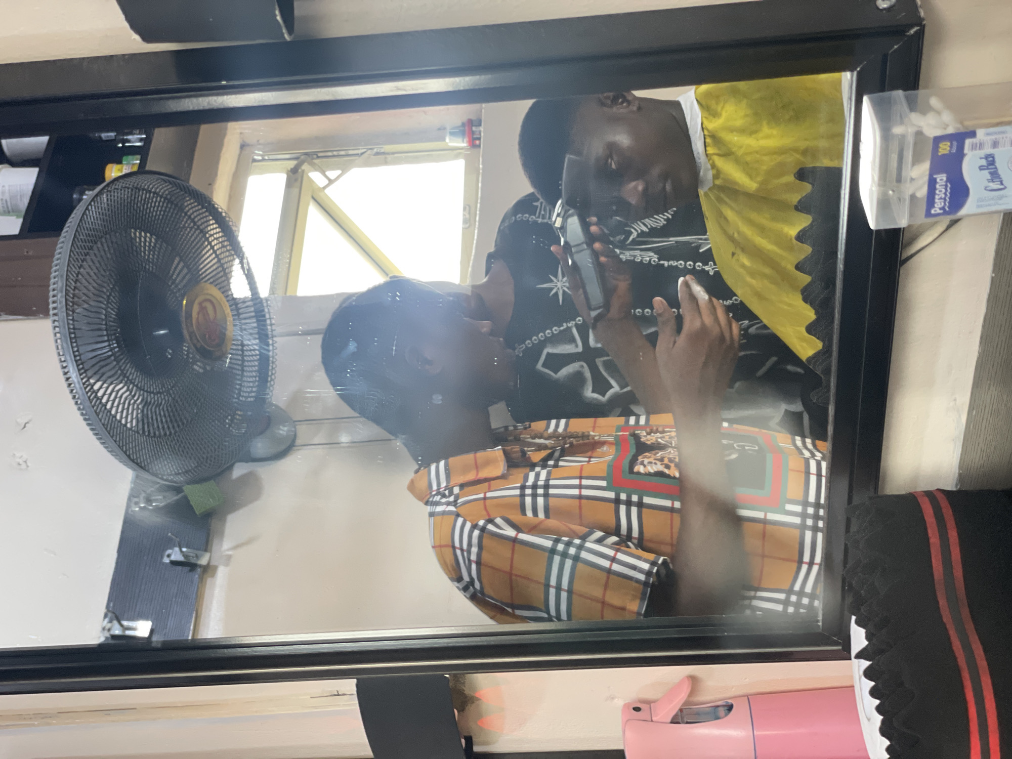 A mirror reflection reveals two dark-skinned men inside a barber shop in Ado, Nigeria. One man is seated wearing a yellow and black shirt, while the other stands, dressed in a plaid shirt. The standing man uses an electric razor to shave the seated man's hair. A black electric fan is mounted on a wall beside the seated man, and a box of cotton swabs sits beside the mirror.