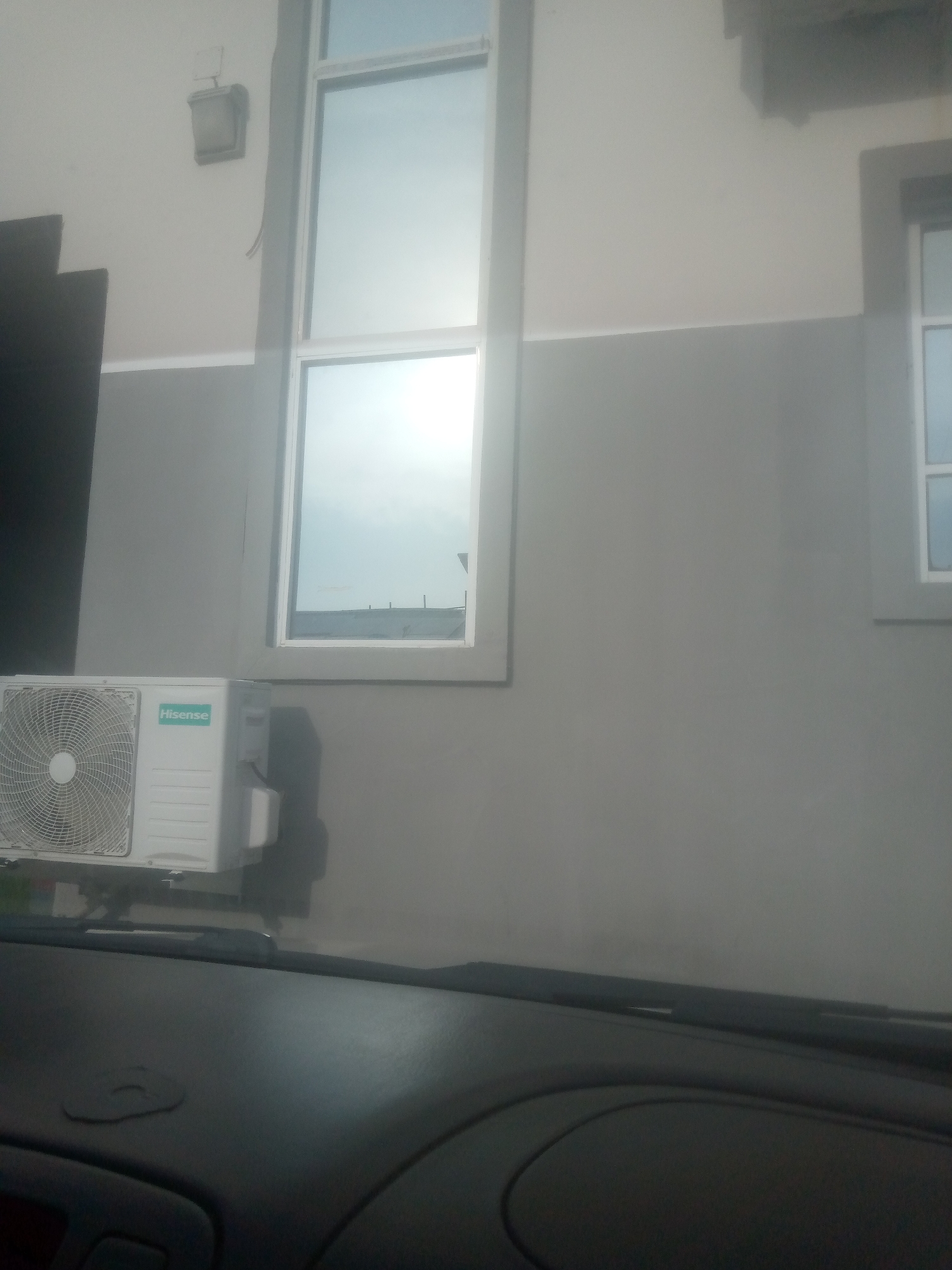 The image shows the exterior of a building with a light gray wall and a white upper section. Two windows are visible, one large, vertical window with a frosted glass pane and a white frame, and a smaller, horizontal window to its right with a similar frame. An outdoor air conditioning unit, labeled "Hisense," is mounted on the wall beneath the large window. The unit is white and has a circular fan grille. The scene appears to be viewed from inside a vehicle, with a dark dashboard and windshield wipers visible in the foreground. This scene is located in Jalingo, Nigeria.
