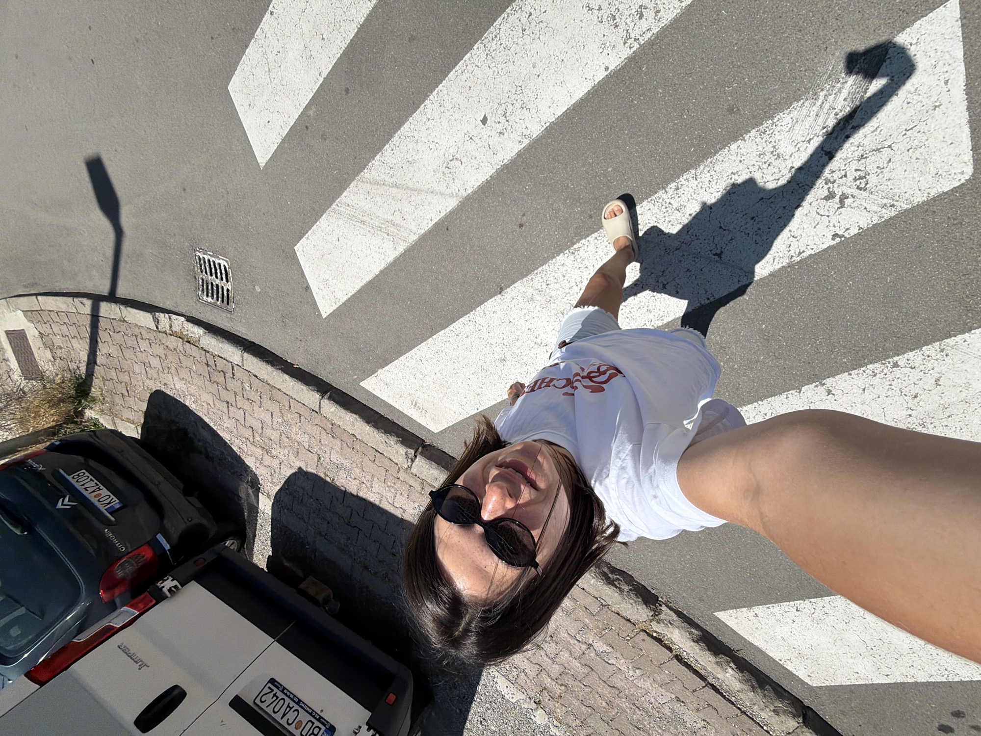 This image is a high-angle selfie taken on a sunny day at a pedestrian crossing in Budva, Montenegro. The primary subject is a person, appearing female, with dark hair and sunglasses, wearing a white t-shirt, light shorts, and light-colored slides. They are positioned on a white stripe of the zebra crossing, extending their right arm upwards to hold the camera for the shot.
The scene depicts a typical urban street environment. To the left, a raised sidewalk is paved with reddish-brown rectangular pavers. Parallel to the sidewalk, a white Citroën Jumper van is parked, displaying the Montenegrin license plate "BD CA042", where "BD" signifies Budva registration. Adjacent to the van's rear is a dark-colored Citroën passenger car (likely a Berlingo or similar model), also parked, with the Montenegrin license plate "KO AZ108", indicating registration in Kotor.
The road surface is asphalt, with prominent white stripes forming the pedestrian crossing. A metal drain grate is visible on the asphalt surface further down the street, near the curb. Long, distinct shadows are cast across the street, including the person's shadow stretching across the crosswalk and the shadow of what appears to be a street lamp or pole. The lighting indicates strong, direct sunlight and a clear sky.