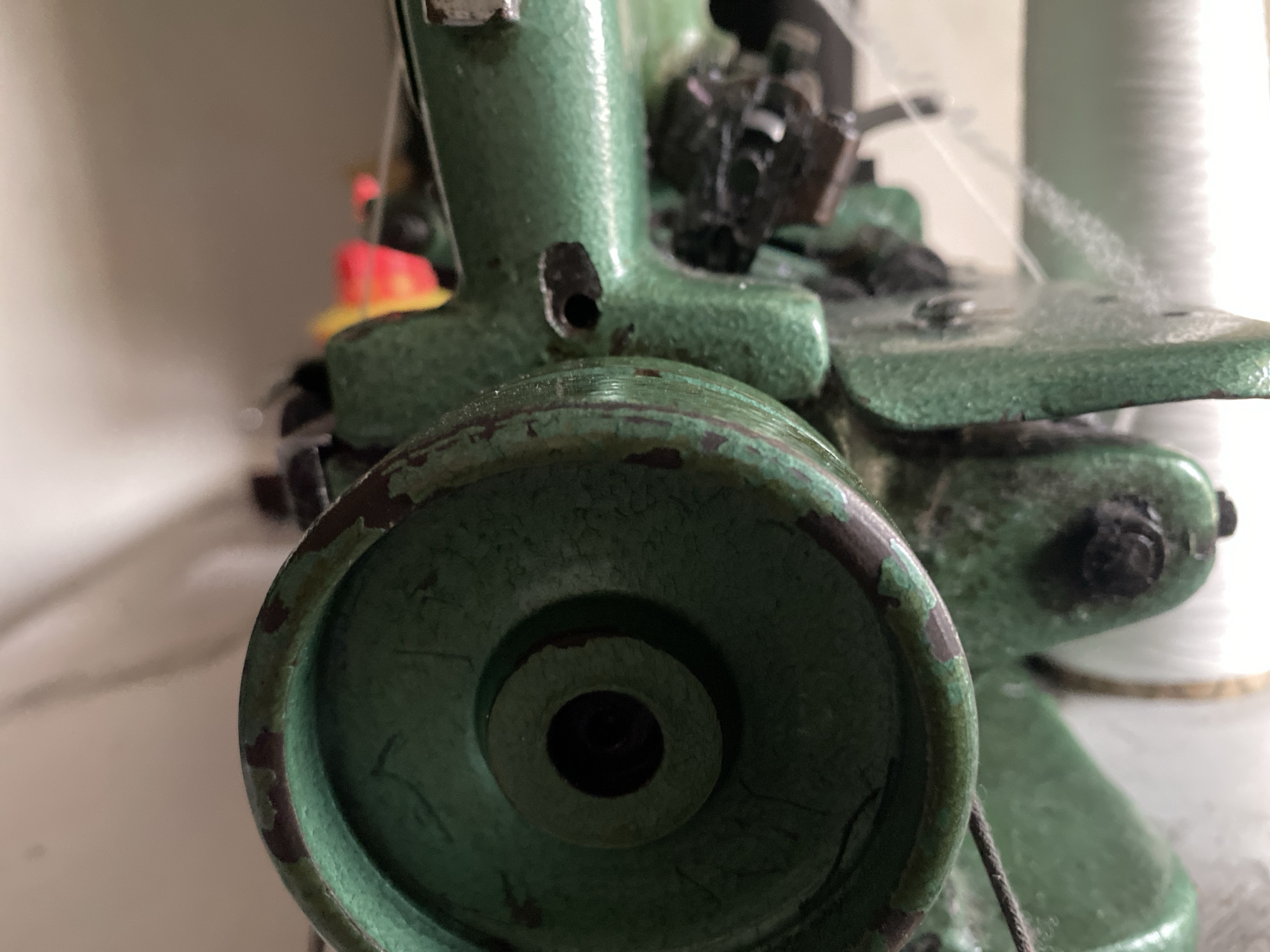 A close-up, low-angle shot focuses on the lower portion of a green industrial sewing machine. The foreground is dominated by a large, circular metal wheel with a weathered, chipped green paint finish, revealing rust underneath. A black circular opening is visible in the center of this wheel. To the right of this wheel, the machine's body, also green, is textured and appears to have some wear. Further back and to the right, a spool of white thread is visible, standing upright. In the background, out of focus, are hints of other objects, including a bright yellow and red element which may be a bobbin or a part of the machine. The overall environment suggests an indoor setting, possibly a workshop or a sewing room. There is no indication of the city of Jauro Yinu, Nigeria, in the image.