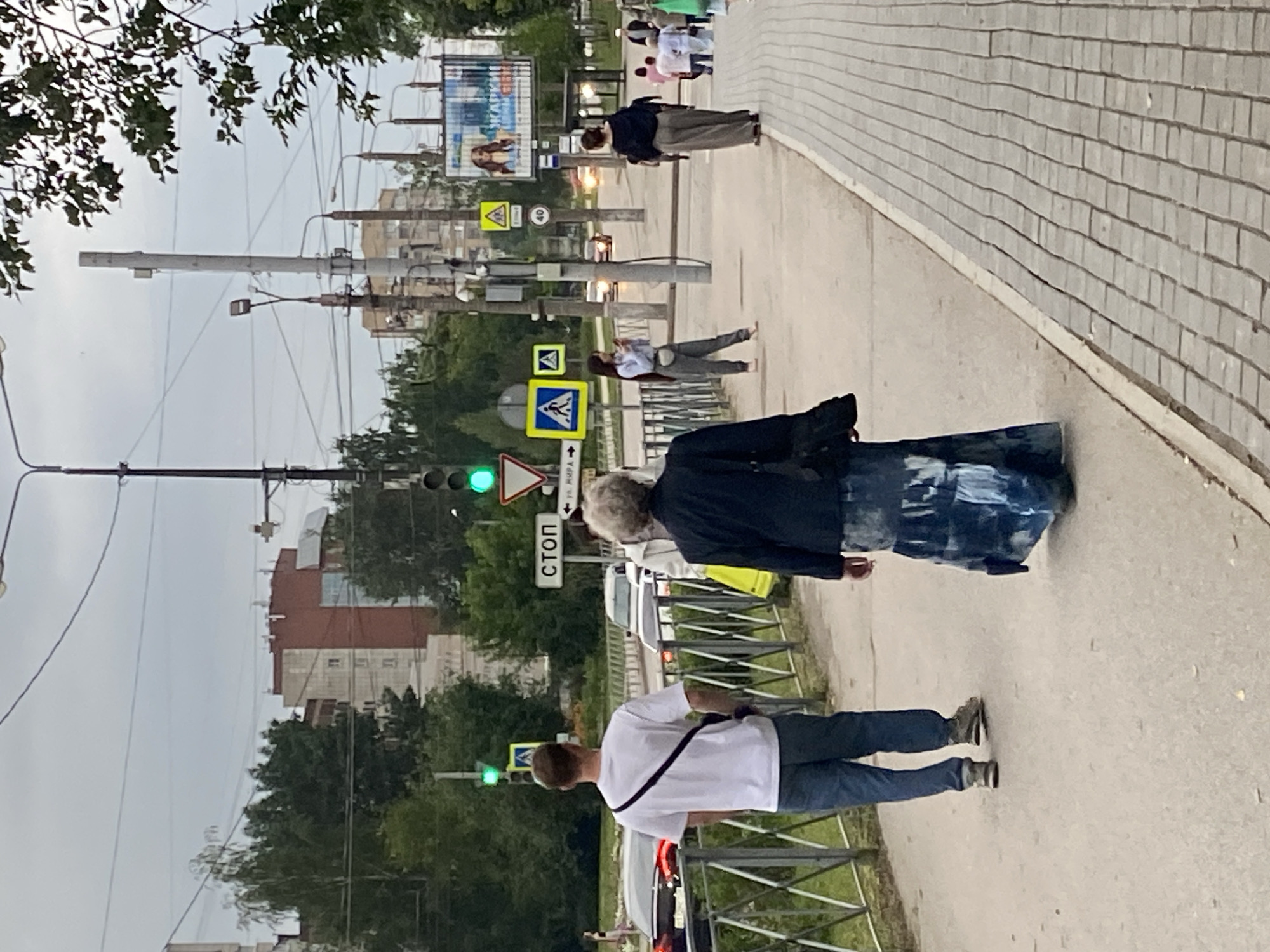 The image depicts a street scene in Perm, Russia, likely on a cloudy day. In the foreground, a person in a white t-shirt and blue jeans walks away from the camera on a paved sidewalk. To their right, an older woman with curly gray hair is also walking away, wearing a dark jacket and a long, patterned skirt.
Behind them, a group of people are visible on the sidewalk and street. Traffic signals show a green light. A "STOP" sign is posted on a pole, indicating a traffic intersection. There are also pedestrian crossing signs. In the background, several buildings and trees line the street, along with streetlights and utility poles. A billboard advertising a product or event is visible on the right side of the image. Cars are present on the road, and a metal fence separates the sidewalk from a grassy area.
