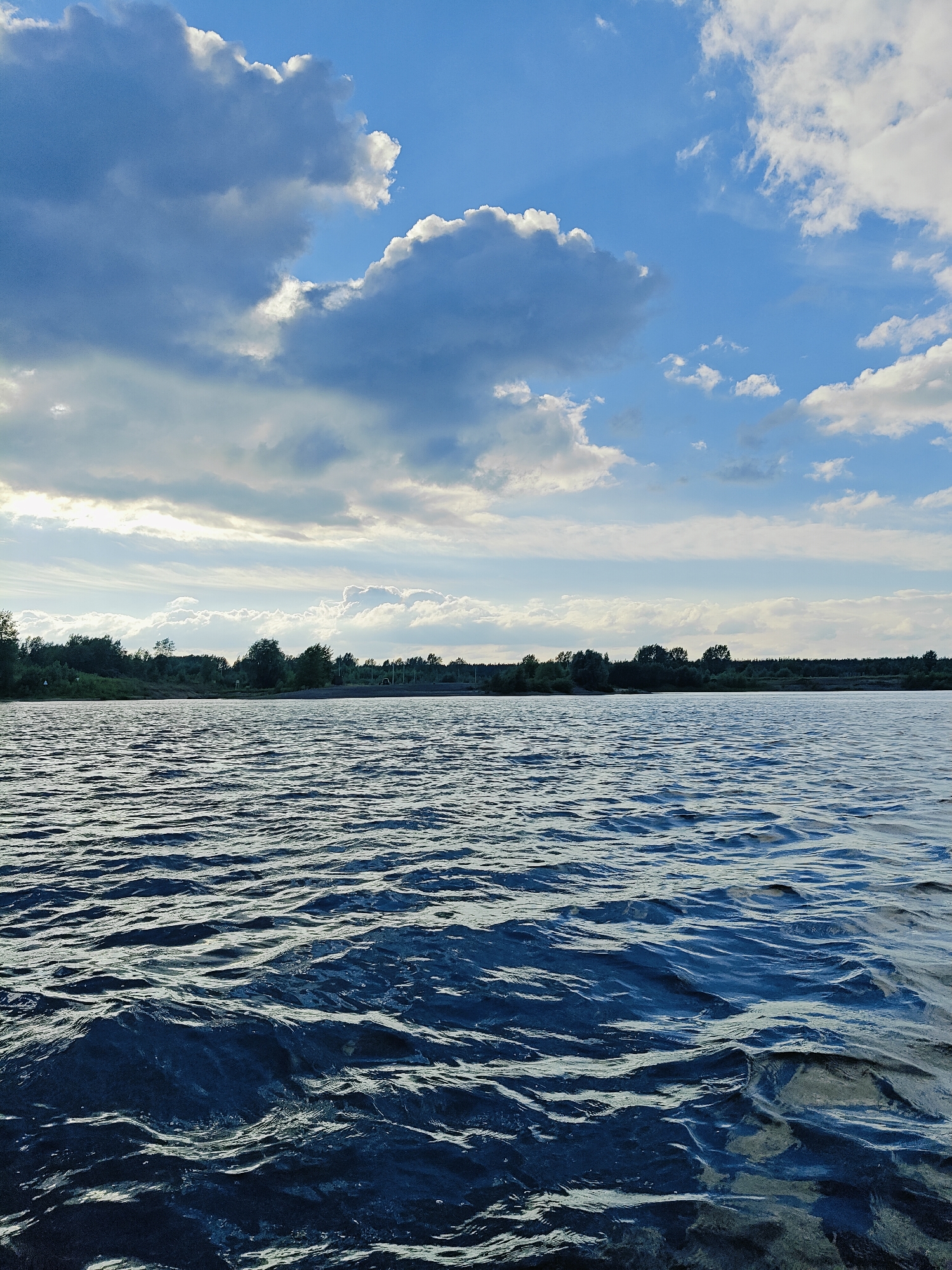 A wide, dark blue body of water with small ripples occupies the foreground and middle ground of the image. Beyond the water, a line of green trees and shrubs forms a dense treeline along the horizon. In the distance, a faint, light-colored structure, possibly a building, is visible behind the trees. The sky above is a bright blue, dotted with a mix of dark gray and white clouds. The overall scene suggests an outdoor natural environment, likely a river or lake, under a partly cloudy sky. No people, animals, or specific urban structures are discernible in the image.