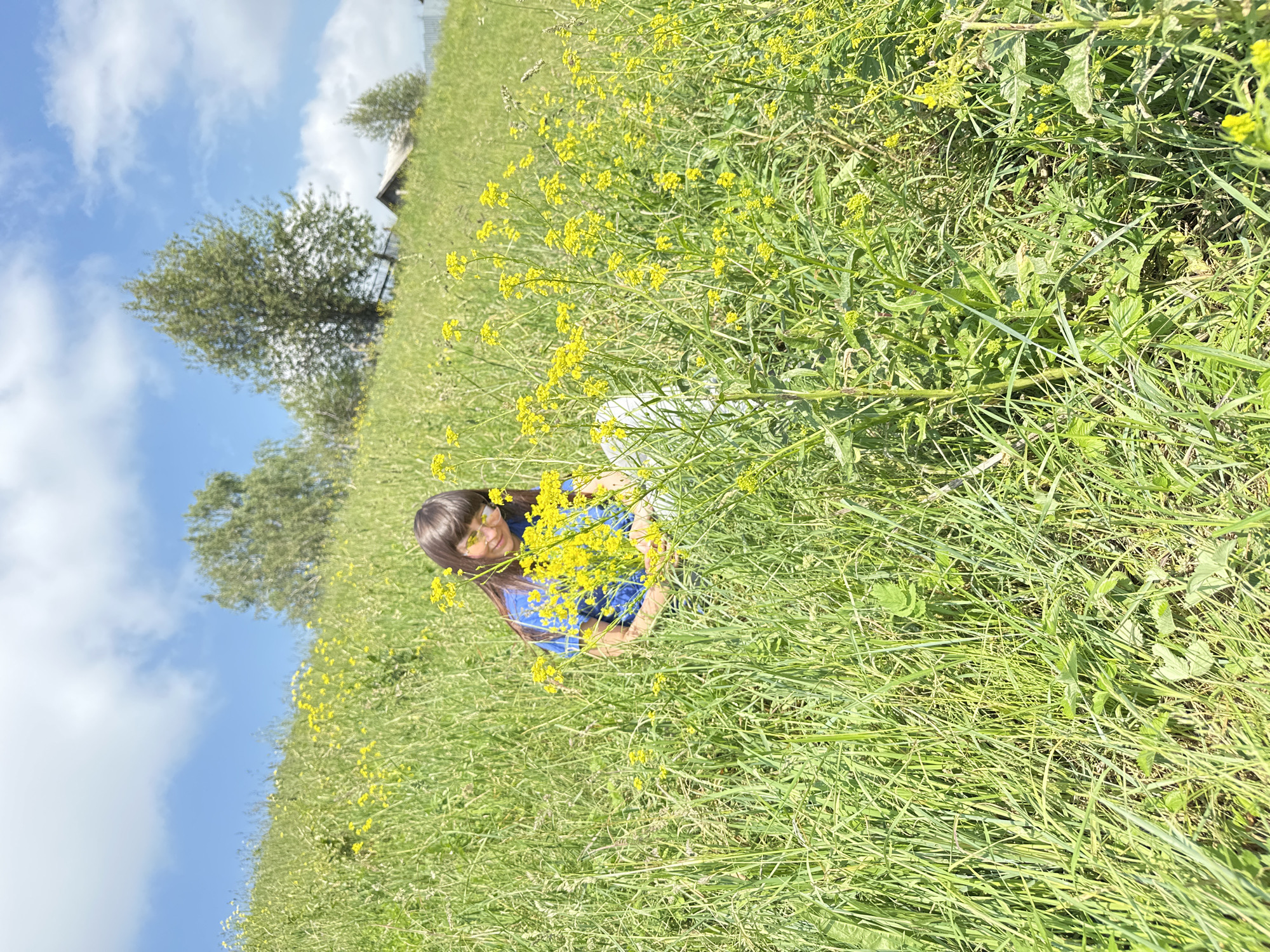 A young person with dark hair and glasses is sitting in a grassy field with yellow wildflowers. The person is wearing a blue shirt and white pants. The field is on a gentle slope, and in the background, there are trees and a rustic wooden building with a thatched roof. The sky is blue with scattered white clouds. The overall impression is one of a peaceful rural scene. The location is the village of Buranovo, Russia.