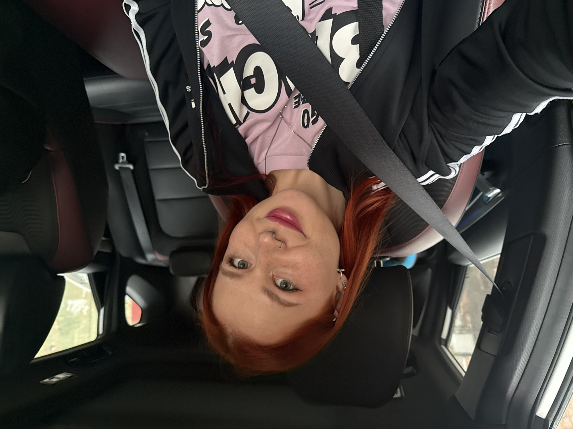 A young, fair-skinned woman with red hair is sitting in the passenger seat of a car, wearing a pink t-shirt with black lettering, a black jacket with white stripes down the sleeves, and a seatbelt. She has freckles, a nose ring, and earrings. The car's interior is black and red. The background shows a blurred view through the car windows, suggesting an outdoor setting with trees and buildings. The location is not identifiable as Rostov-on-Don, Russia, based on the provided image.