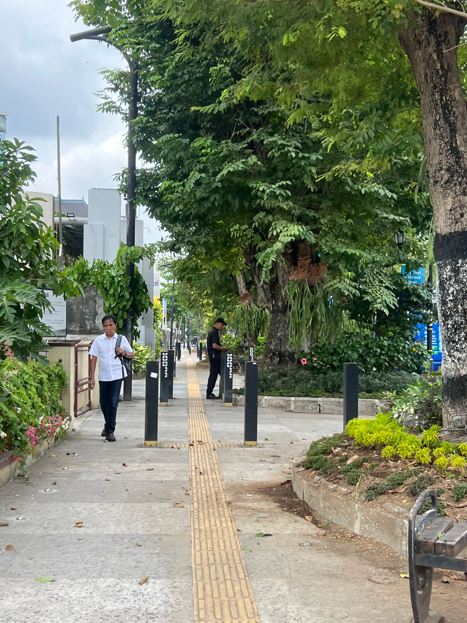 A man in a white short-sleeved shirt and dark pants walks on a paved sidewalk in Yogyakarta, Indonesia. He carries a black bag over his left shoulder. A second man in a black uniform stands further down the sidewalk, facing away from the camera. The sidewalk is lined with black bollards and has a tactile paving strip. Lush green trees and foliage line the sidewalk, with some flowering plants visible on the left. Buildings can be seen in the background. The scene appears to be a public walkway in a city park or a well-maintained street.