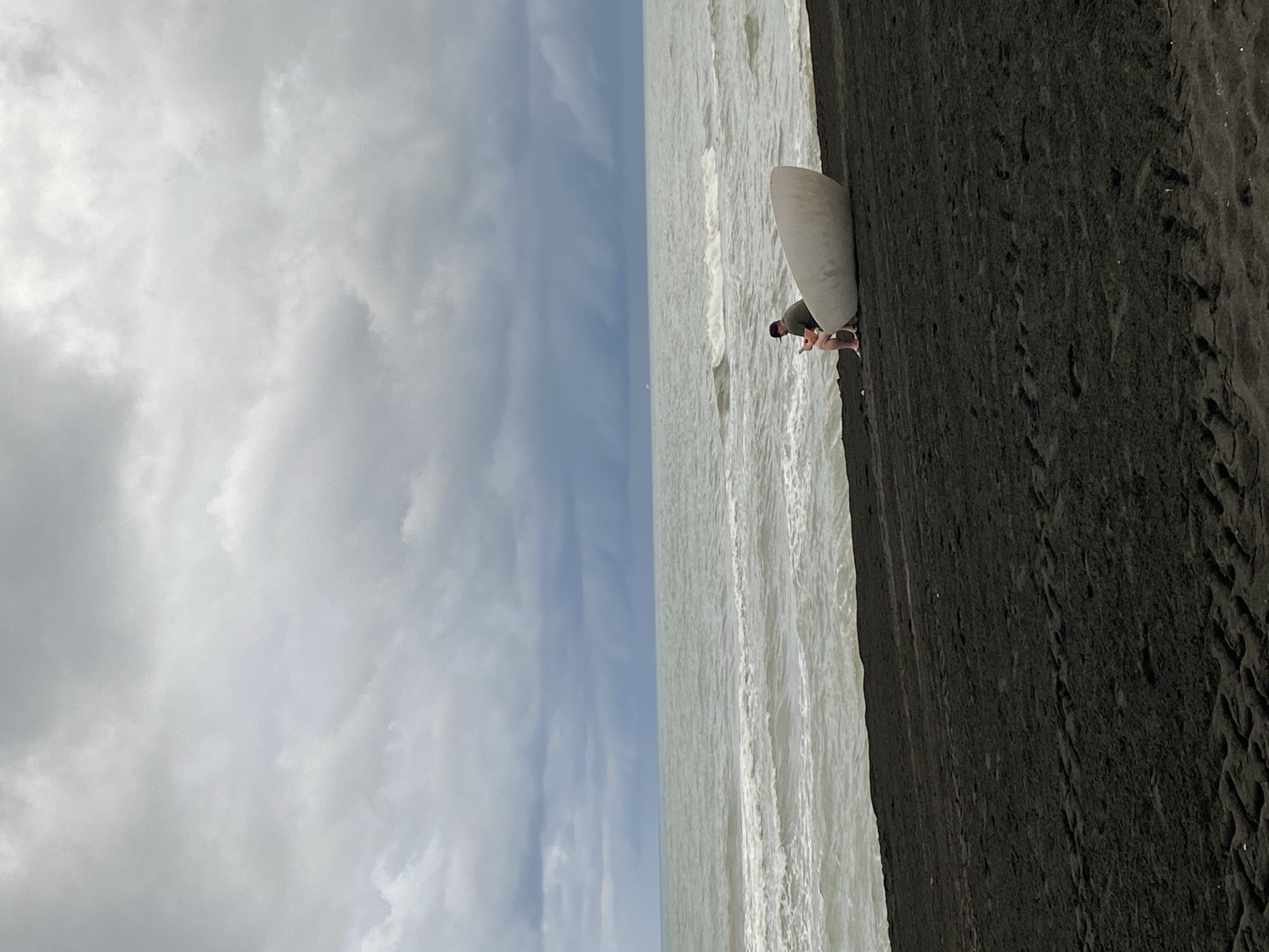 A solitary individual sits on a black sand beach in Ureki, Georgia, next to a white, boat-shaped object. The person, wearing a dark green t-shirt and shorts, is looking down at a mobile device. The sea in the background is a muted grey with moderate waves, and the sky is heavily overcast with dark, dramatic clouds. The black sand beach is marked with tire tracks, suggesting vehicle activity.