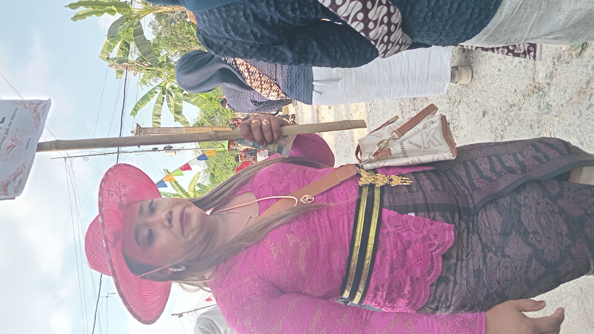 A woman in a bright pink lace kebaya and a patterned brown sarong stands holding a bamboo pole. She wears a wide-brimmed pink hat and a tan crossbody bag with brown trim. The background shows a street scene in Kedungwaru, Indonesia, with banana trees, flags, and other people in traditional clothing. The sky is bright and overcast.