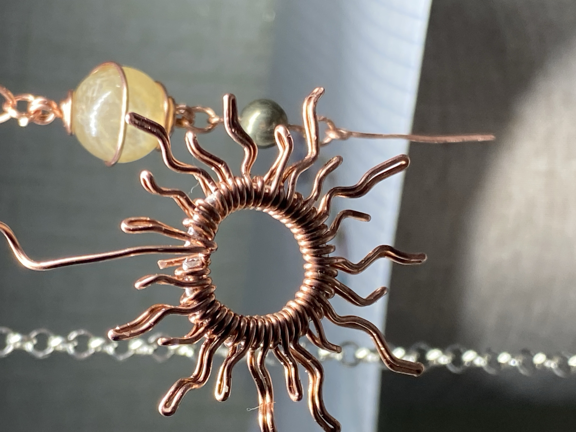 The image displays a close-up, macro photograph of a copper wire-wrapped sun pendant, suspended alongside a silver chain. The pendant is intricate, with a circular core formed by tightly wound copper wire and numerous radiating spikes, also crafted from copper wire, that are curved and wavy. To the upper right of the sun pendant, a small, translucent yellow bead is attached to a copper chain. Below this bead and to the right of the sun pendant, a small, metallic grey, spherical bead is suspended. The background is softly blurred, with hints of light and shadow suggesting an indoor setting, possibly a workshop or a display. There are no people or animals visible in the image, nor are there any discernible elements that would place the scene in Kushchovskaya, Russia. The focus is entirely on the handcrafted jewelry.