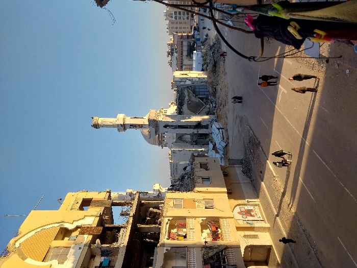 The image portrays a war-torn city landscape. Heavily damaged buildings stand next to a partly destroyed mosque. A street runs through the scene, with several people walking along it. Clothes hang on a line in the foreground. The overall atmosphere evokes devastation and resilience in the face of destruction. The sky is clear and blue.