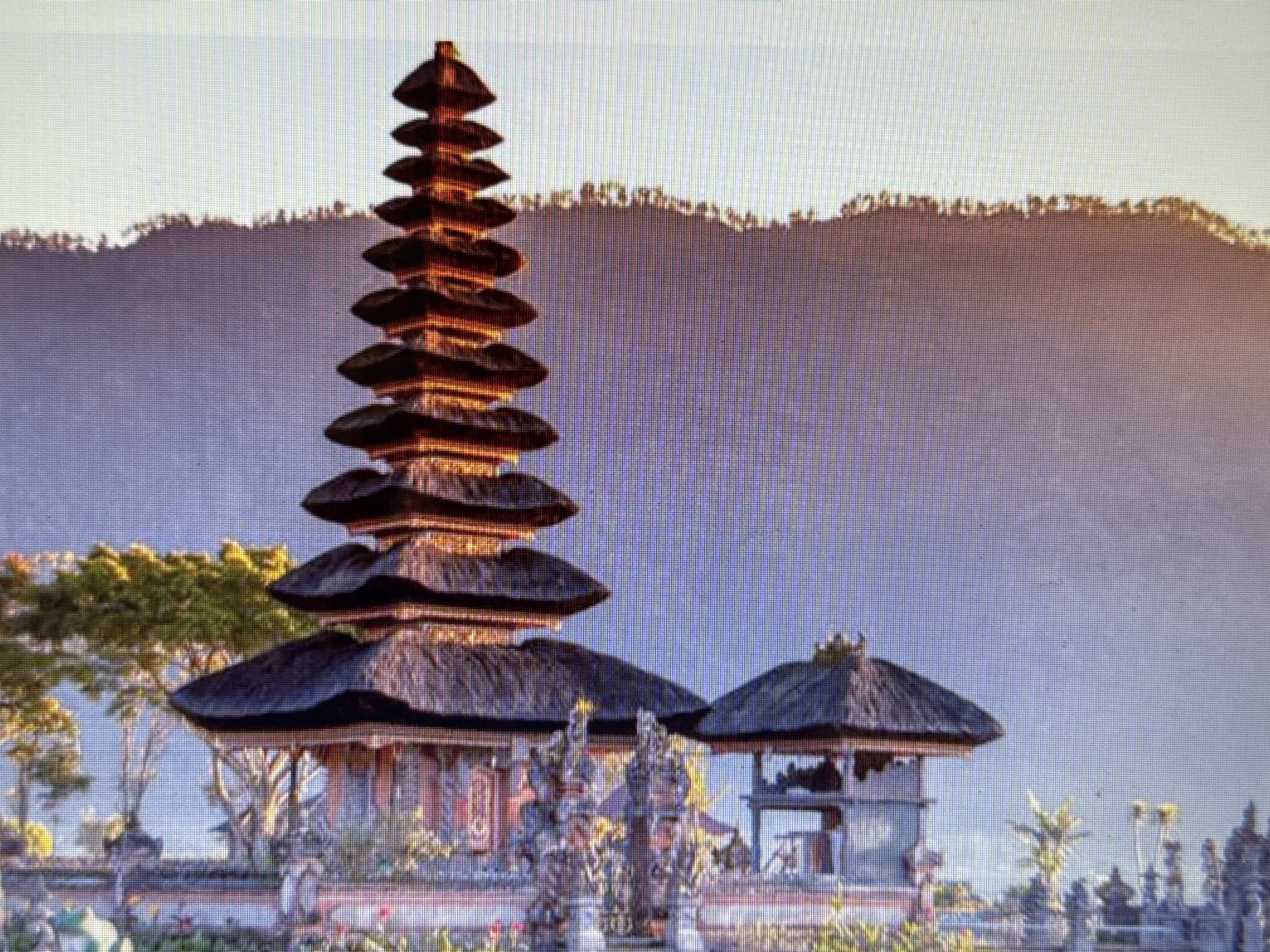 The image depicts a serene landscape featuring a multi-tiered traditional temple structure. The temple, with its dark, thatched roofs and intricate stonework, is prominently positioned on the right side of the frame. It is illuminated by warm, golden light, suggesting it might be sunrise or sunset.
Behind the temple, a lush green tree stands tall, its branches reaching towards the sky. In the background, a hazy, purple mountain range forms a dramatic backdrop, its silhouette softened by the distance and possibly a light fog or mist. The sky above transitions from a pale, almost white hue at the horizon to a soft blue further up.
The foreground shows hints of a garden or landscaped area with various plants and decorative elements, including what appear to be smaller statues or ornamental pillars. The overall scene evokes a sense of tranquility and cultural significance. The specific location cues in the image do not indicate Pushkino, Russia, as the architecture and landscape are more characteristic of Southeast Asian settings, likely Indonesia or a similar region. There is no visible text in the image.