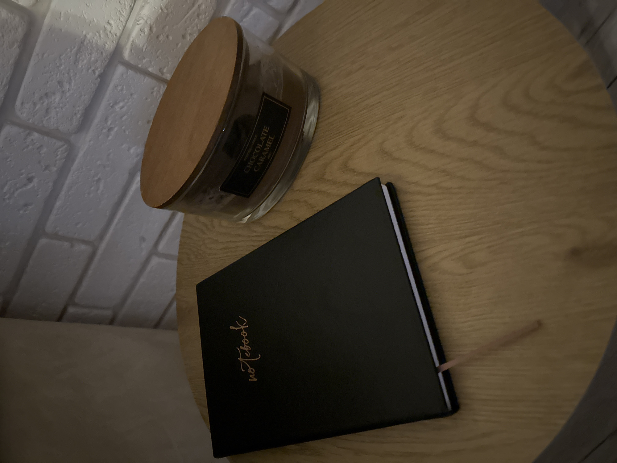 The image shows a wooden side table with a black notebook, a "Chocolate Caramel" scented candle, and a pale brick wall in the background. The notebook has "Notebook" written on the cover in cursive. The table appears to be in a room with indirect lighting. No people are present.