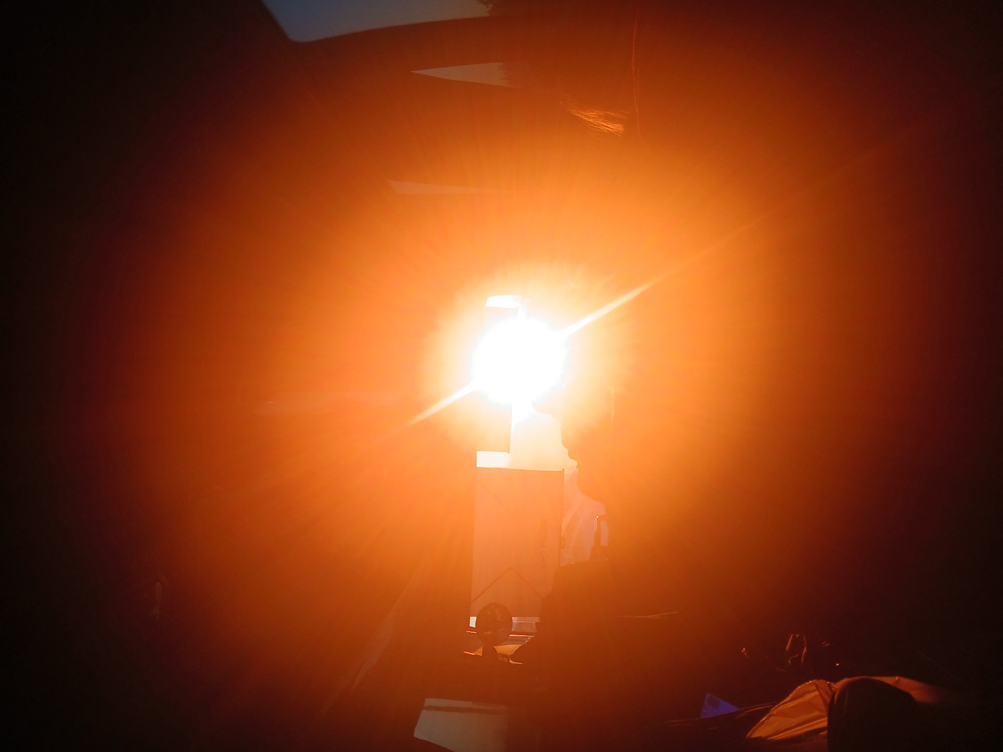 The image depicts an indoor scene dominated by a bright, concentrated light source. The light is orange-tinted and obscures much of the surroundings. Silhouetted forms of objects are visible in the foreground and midground. A tall rectangular shape can be discerned in the center, possibly a box or a screen, directly in front of the light. To the right of the light source, the vague outline of a person's head and shoulders is visible, also silhouetted. The background is dark and indistinct. No elements indicate that the location is Ustinovo, Russia.