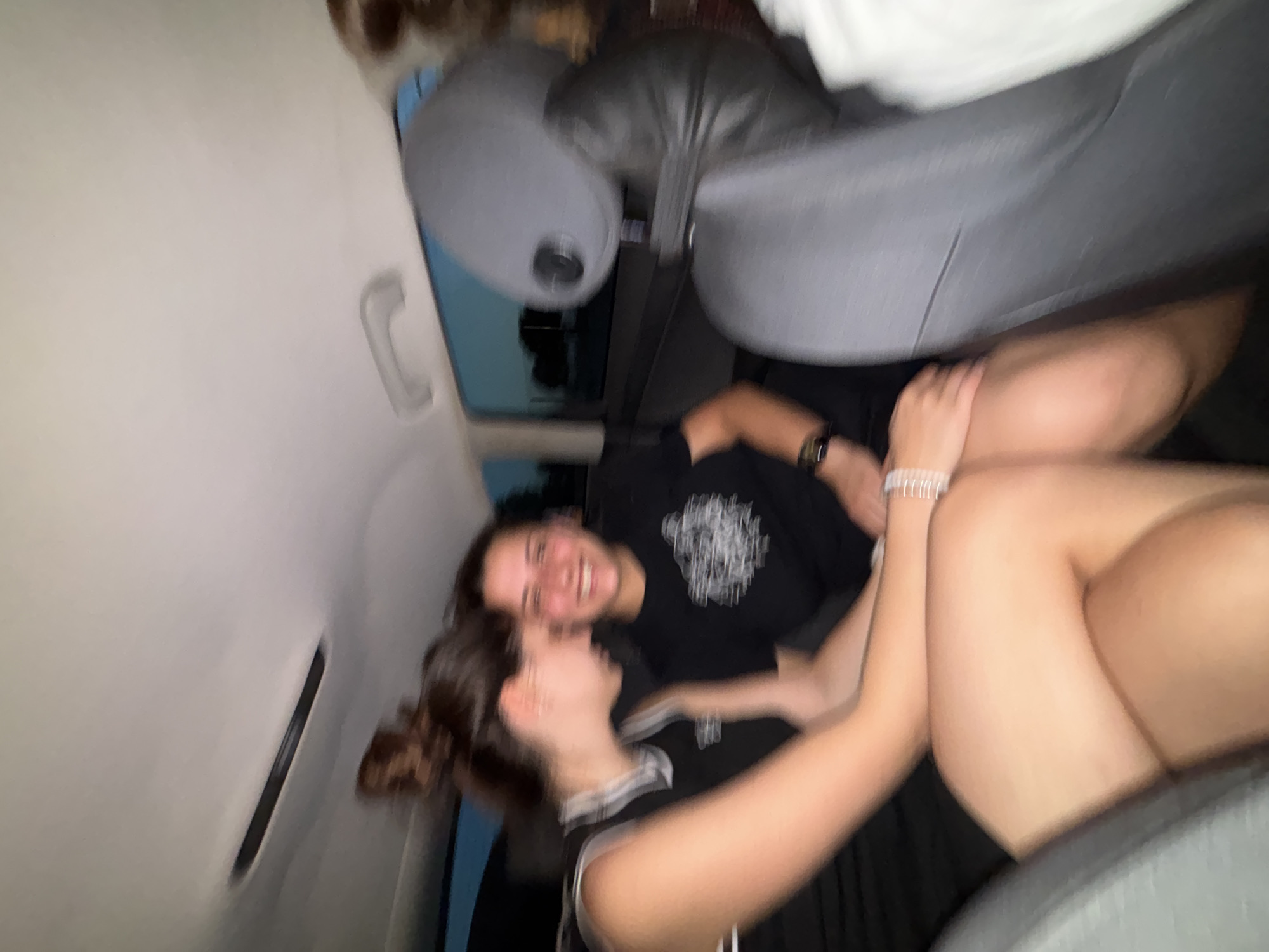 Two individuals are seated in the backseat of a vehicle. The person on the left, wearing a black and white athletic top, has their head tilted down. The person on the right, wearing a black t-shirt with a graphic print, is smiling and looking towards the viewer. The interior of the car is visible, including headrests and a window showing an outdoor scene. The image appears to be taken at dusk or in low light conditions. The scene is located in the city of Mramor, Bulgaria.