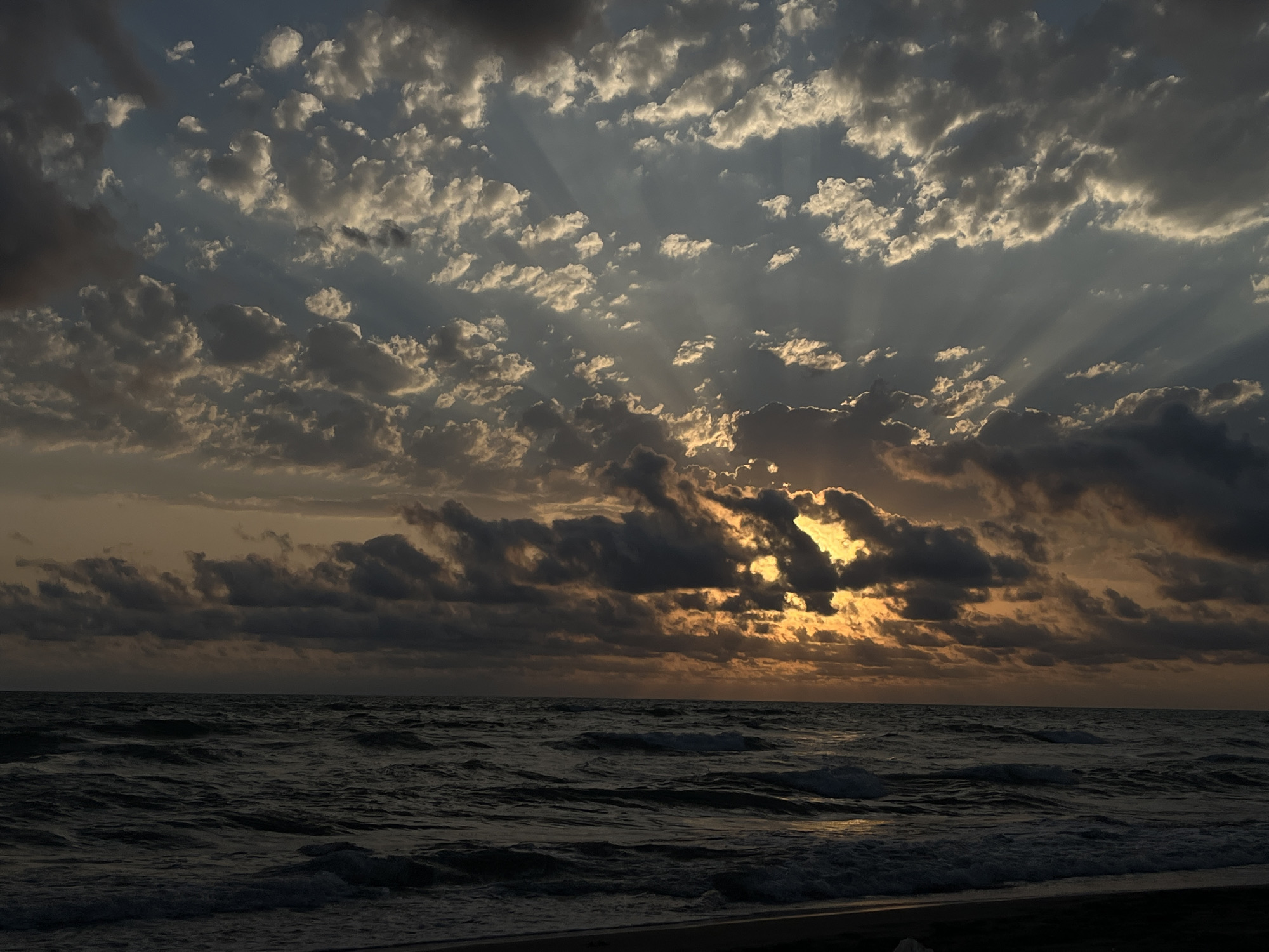 The image depicts a dramatic sunset over the Black Sea, viewed from the coast of Supsa, Georgia. Dark, heavy clouds dominate the sky, interspersed with patches of brighter sky where the sun's rays are breaking through, creating a crepuscular effect. The sea is rough, with visible waves and whitecaps, reflecting the subdued light of the sunset. The overall impression is one of atmospheric intensity and natural beauty. There are no people or man-made objects visible in the frame.