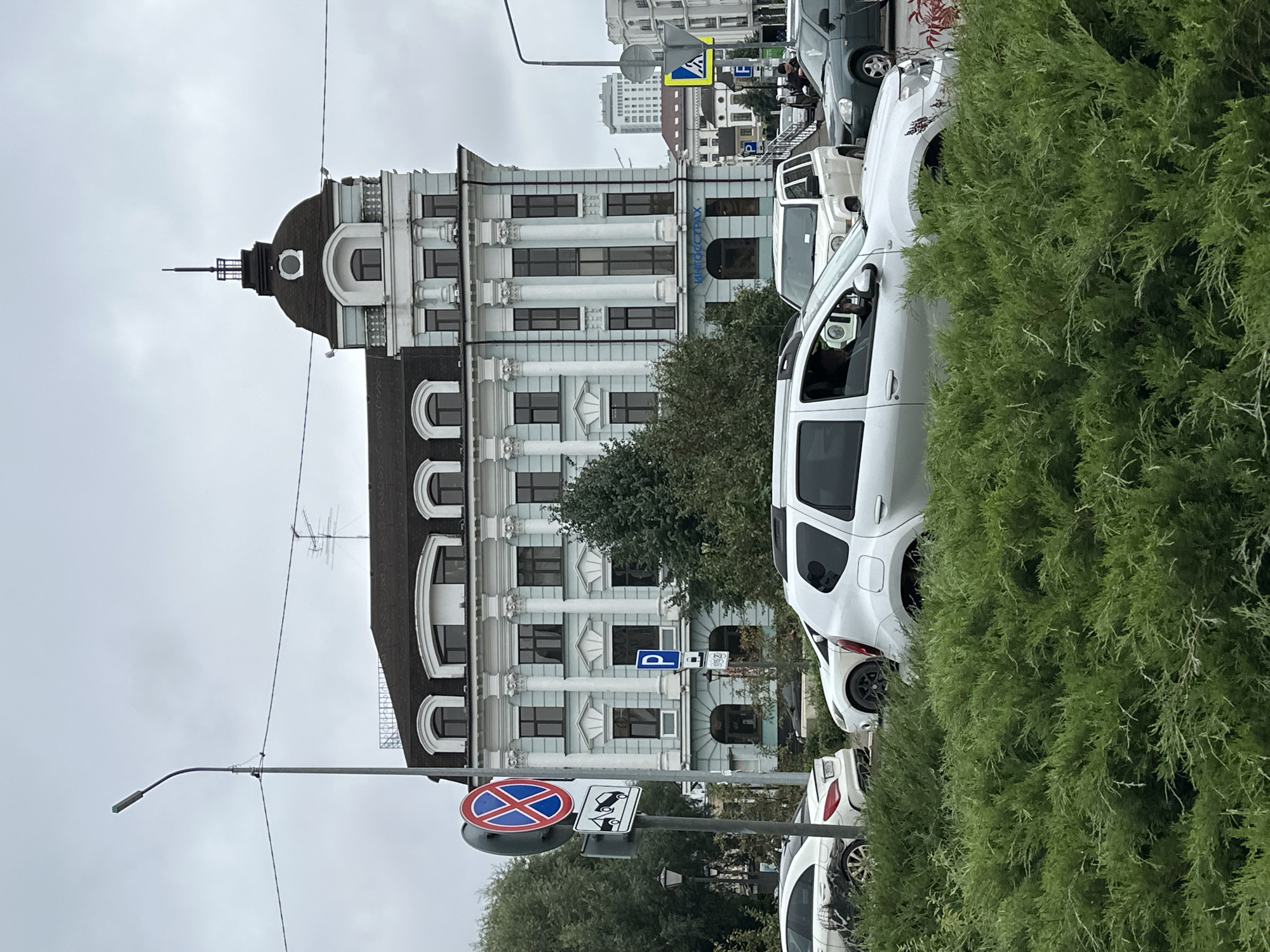 The image depicts a street scene in Kazan, Russia, with a prominent light blue building featuring classical architectural elements, including columns and arched windows, dominating the background. A dark domed roof with a spire caps the building. In the foreground, a dense evergreen bush partially obscures several vehicles, including a silver Dacia Duster, a white Mercedes-Benz, and a white SUV. Traffic lights and various road signs, including a "no stopping" sign and a parking sign, are visible. The sky is overcast, indicating a cloudy day. The building in the background has Cyrillic lettering on its facade, which appears to read "ИНГОССТРАХ" (INGOSSTRAKH), a Russian insurance company. The overall impression is that of a typical urban environment in Russia.