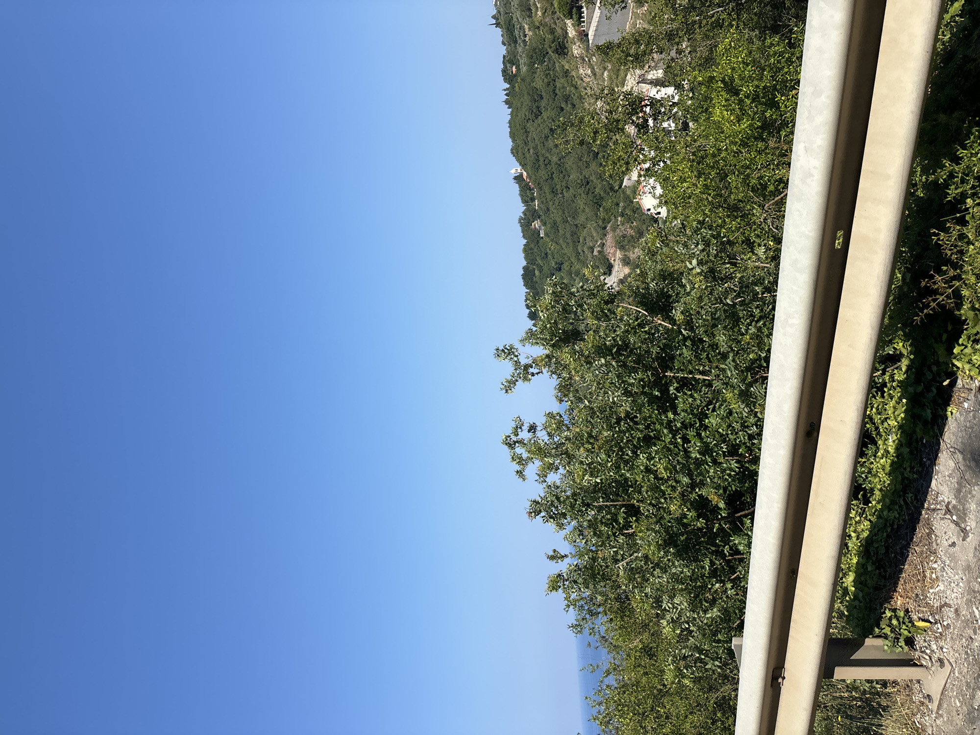 The image depicts a view from a road overlooking a lush, green mountainside in Gornji Katun, Montenegro. The scene is dominated by a bright blue sky and the dense foliage of trees covering the sloping terrain. The road itself is visible in the foreground, with a sturdy metal guardrail running along its edge. The guardrail appears to be made of light-colored metal, possibly aluminum or galvanized steel, and shows signs of wear and tear.
Beyond the trees, scattered buildings with white or light-colored roofs are visible, suggesting a small village or scattered houses nestled into the mountainside. In the distance, a body of water, likely the sea, can be seen reflecting the blue of the sky, indicating a coastal or near-coastal location. A slender white structure, possibly a church tower, rises above the trees on the hillside, adding a point of interest to the landscape.
The lighting suggests it is daytime, with clear, sunny weather. The shadows cast by the guardrail and the foliage indicate that the sun is high in the sky. The overall impression is one of a serene and scenic rural environment. There is no visible text in the image. The camera appears to be tilted, giving a slightly angled perspective on the scene.