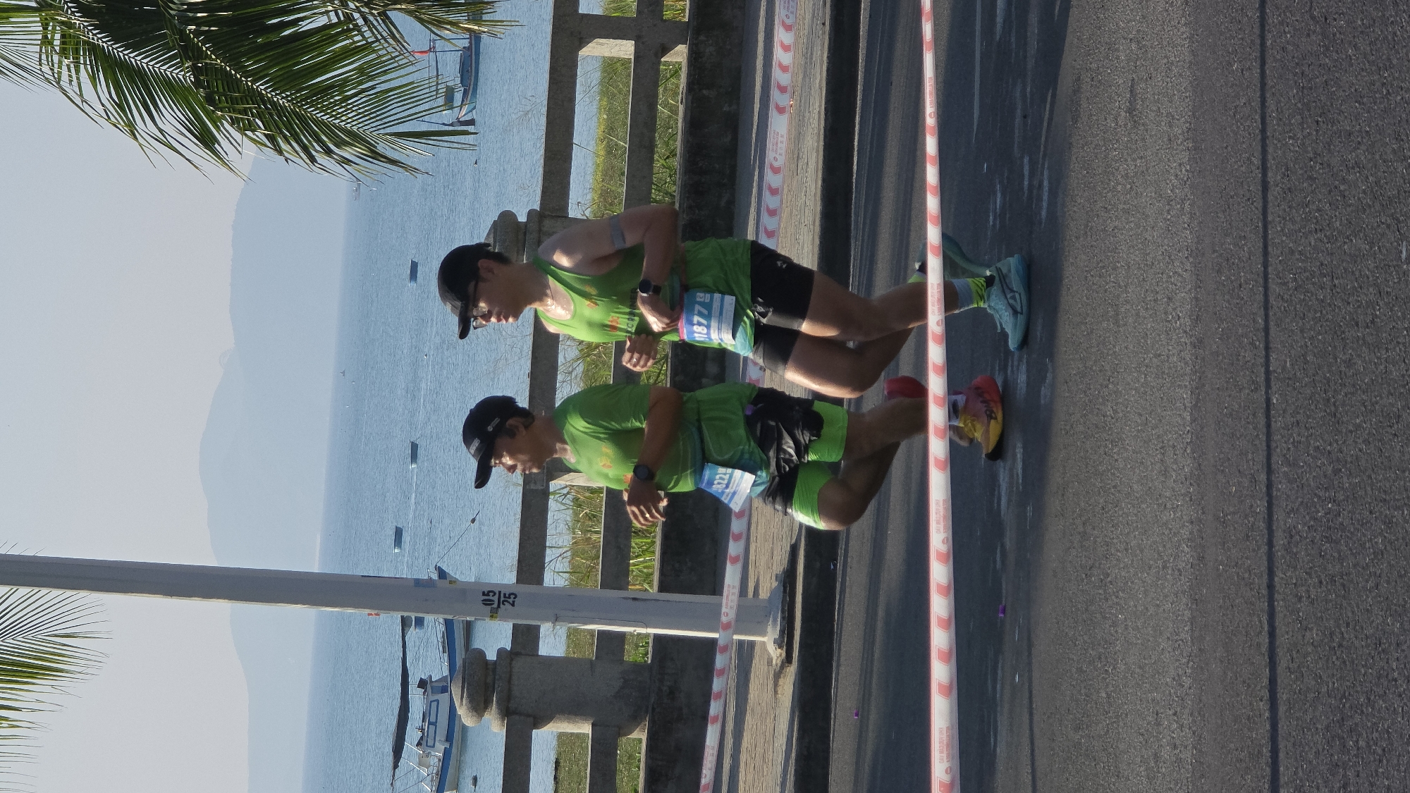 Two men wearing green athletic shirts, black shorts, and baseball caps are running side by side on a paved road along a waterfront. The man in the foreground has a race number "4322" and the man behind him has a race number "1877". There is a red and white striped barrier to their right, separating them from the road. To their left, a concrete barrier lines the edge of a walkway with a view of the ocean, mountains, and several boats. A palm tree is visible in the upper left corner. The scene appears to be in Nha Trang, Vietnam, given the background and coastal setting.