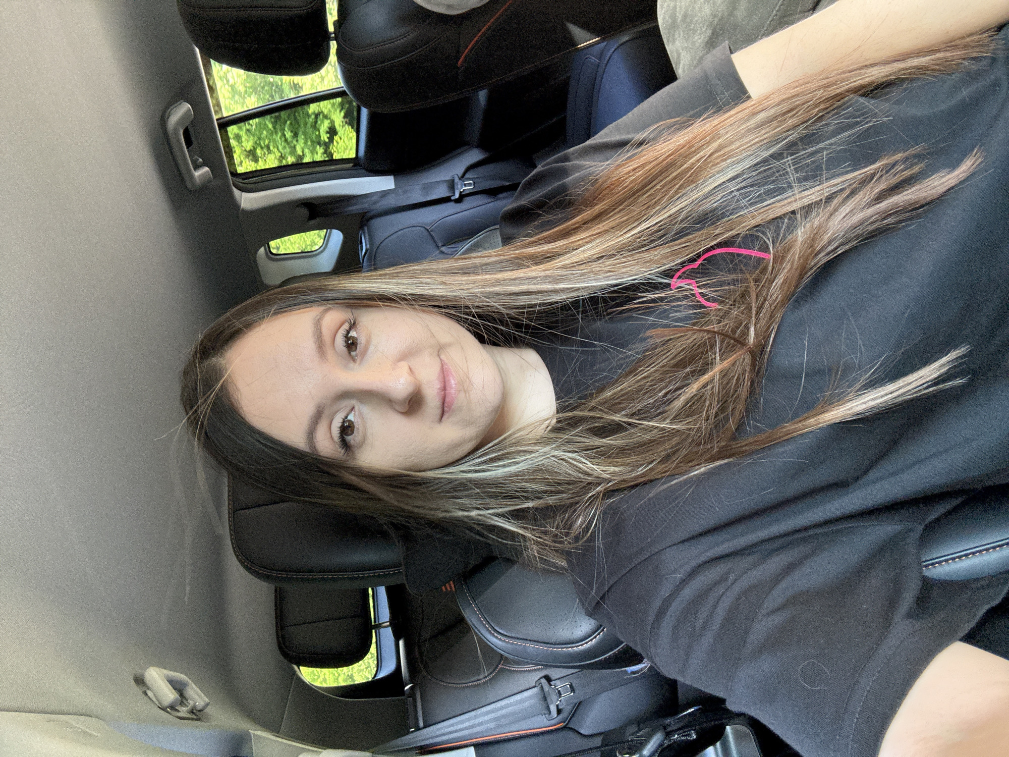 A young woman with long, dark, and blonde-streaked hair is sitting in the driver's seat of a car, looking towards the camera. She is wearing a black t-shirt with a pink heart graphic on the left chest. The interior of the car is visible, with black leather seats and seatbelts. Sunlight filters in through the windows, illuminating the green foliage outside. The woman's pose is casual, and she appears to be taking a selfie. The scene is set during the day, likely in a suburban or rural area, given the presence of trees outside the car. No specific landmarks or indicators place the location in Kroyanskoye, Russia.