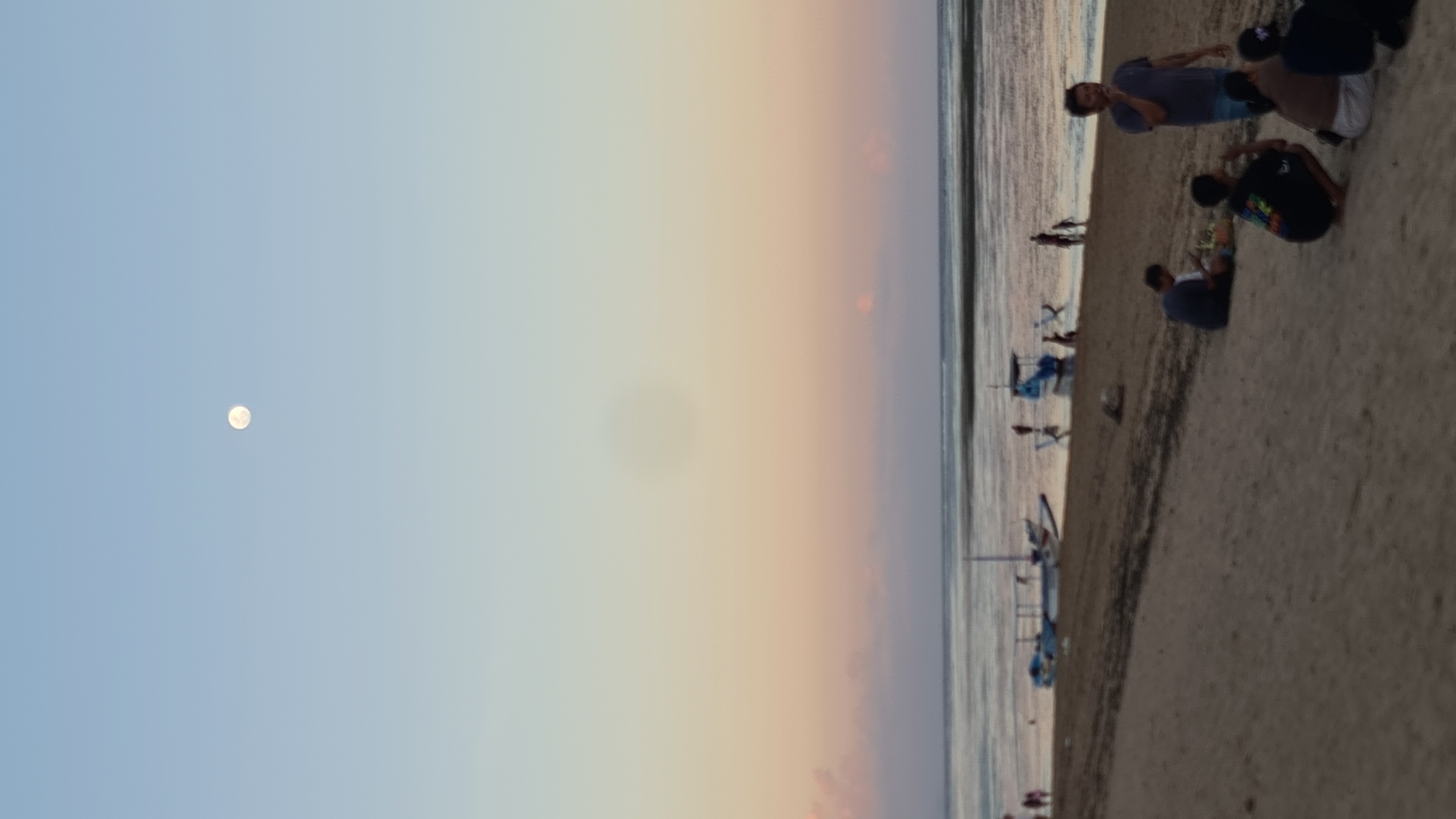 In Denpasar, Indonesia, at dusk, the sky exhibits a gradient of soft blues and pale oranges, with the full moon prominently visible in the upper portion of the frame. The scene unfolds on a sandy beach adjacent to a calm body of water. Several traditional Balinese fishing boats, known as jukungs, are positioned near the shore, some resting on the sand and others partially submerged.
On the beach, groups of people are scattered, engaged in various activities. In the foreground, a man stands talking, while several individuals are seated on the sand, some looking at their phones, others in conversation. Further down the beach, near the water's edge, more people are visible, some standing and others interacting near the boats. The overall atmosphere suggests a tranquil evening gathering by the sea.