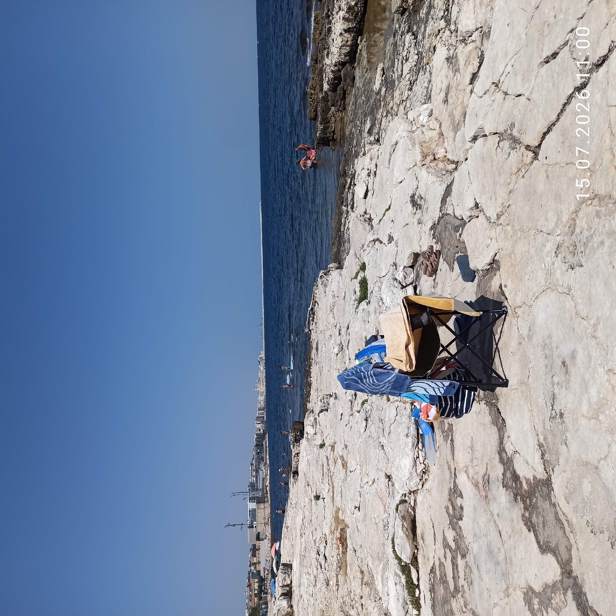 The image shows a rocky shoreline on a clear day, with the city of Molfetta, Italy, visible in the background. The foreground is dominated by a black folding chair with several towels draped over it, suggesting a beach setup. There are also flip-flops and a pair of blue swim fins near the chair. The sea is a deep blue, with a few people visible swimming or wading in the water. The city in the distance features buildings with white facades and some industrial structures, including cranes and tall poles. The overall impression is a sunny day at a coastal location.