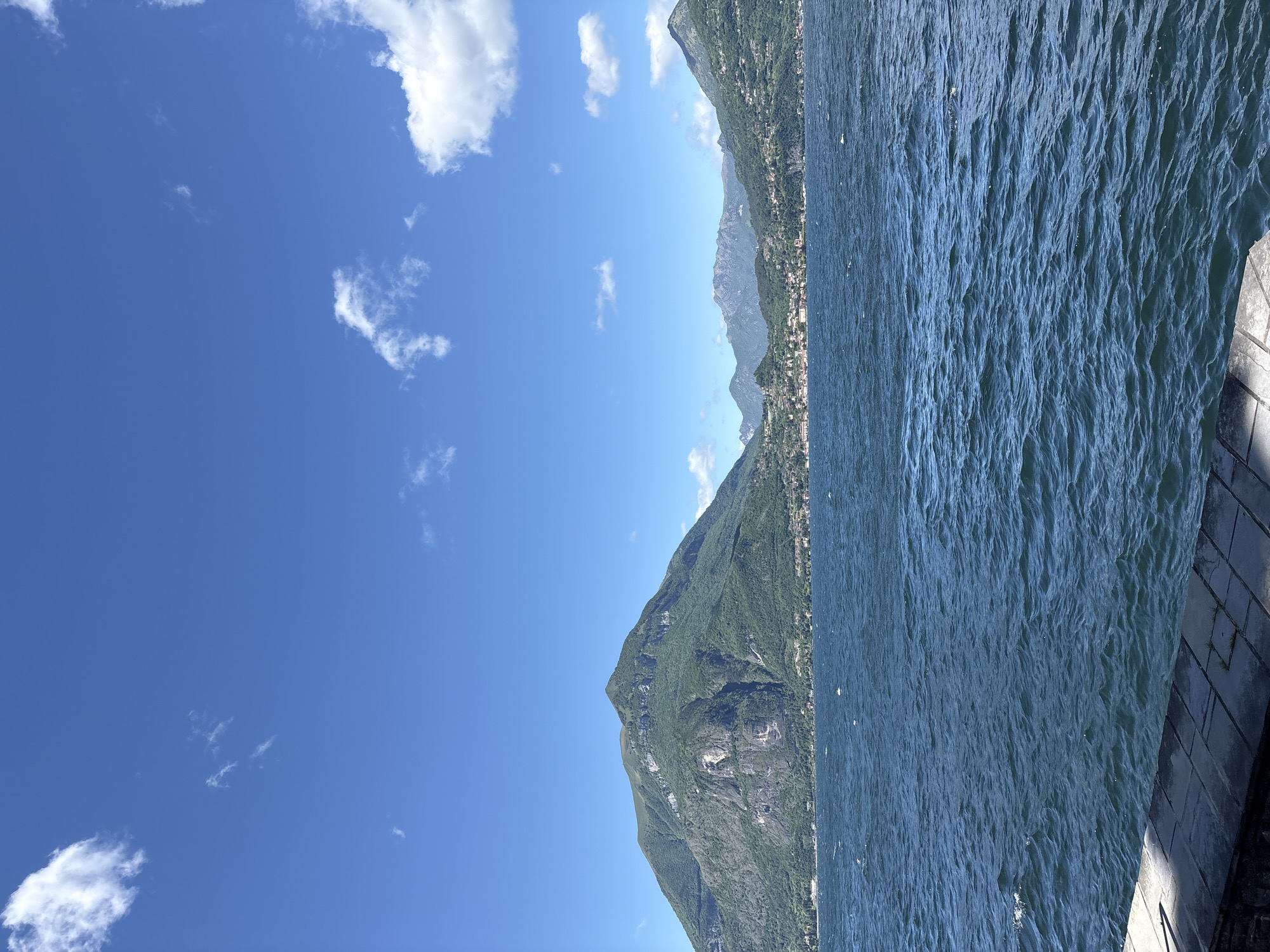 The photograph captures a scenic view in Varenna, Italy. In the foreground, turbulent water fills a significant portion of the frame. The water meets a stone embankment along the right edge of the frame.
Across the water are verdant mountains ascending to the horizon. A cluster of buildings dots the base of one of these mountains.
Above the mountains, the sky is blue, marked by sparse white clouds.